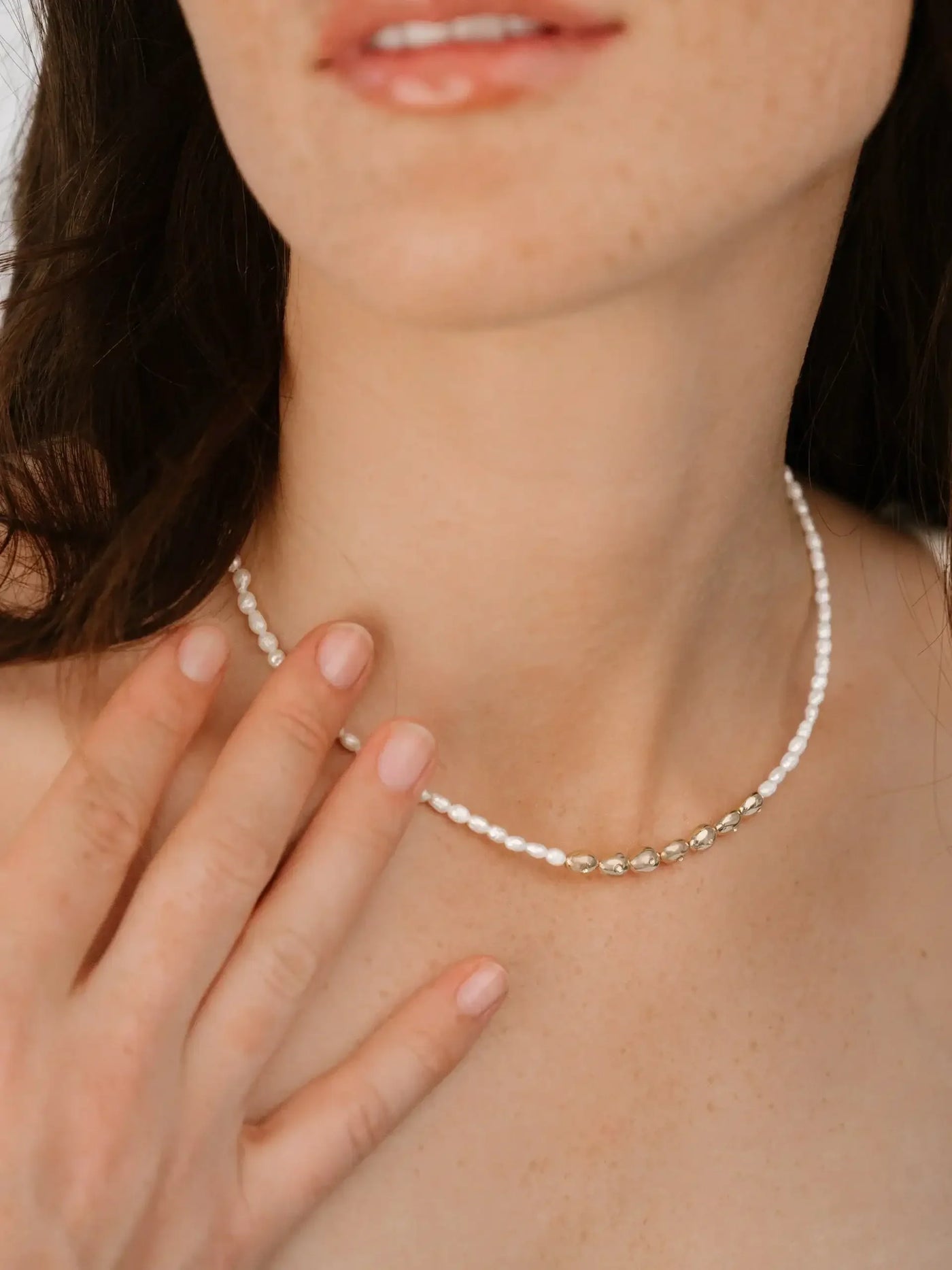 A woman gently touches a delicate necklace made of small pearls and golden accents. The context is intimate, with focus on her bare neck and subtle facial features.