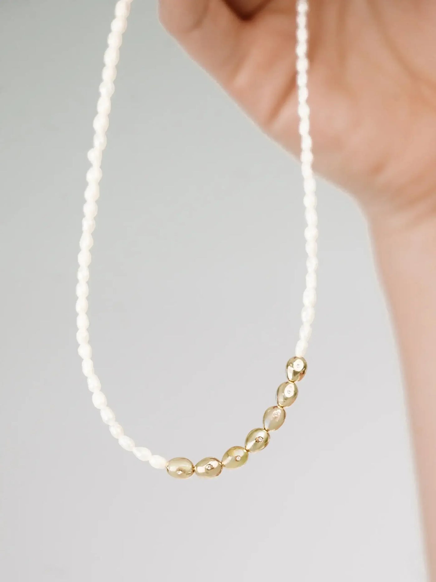 A hand holds a delicate necklace featuring alternating strands of small white pearls and shiny gold beads, displayed against a light gray background.