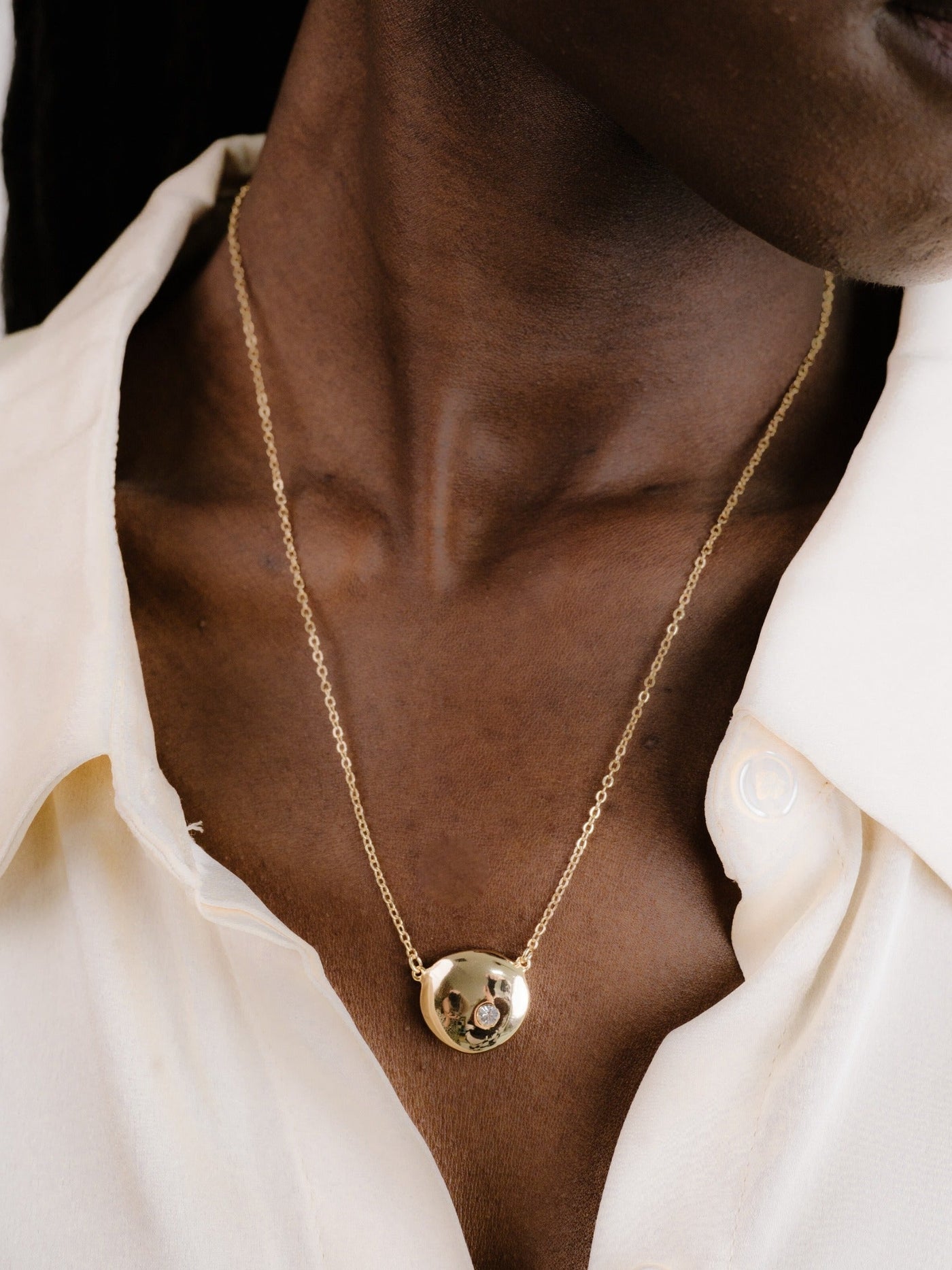 A gold necklace featuring a round pendant with a small gemstone hangs around a person's neck, partially visible above a white, collared shirt, emphasizing a stylish accessory against dark skin.
