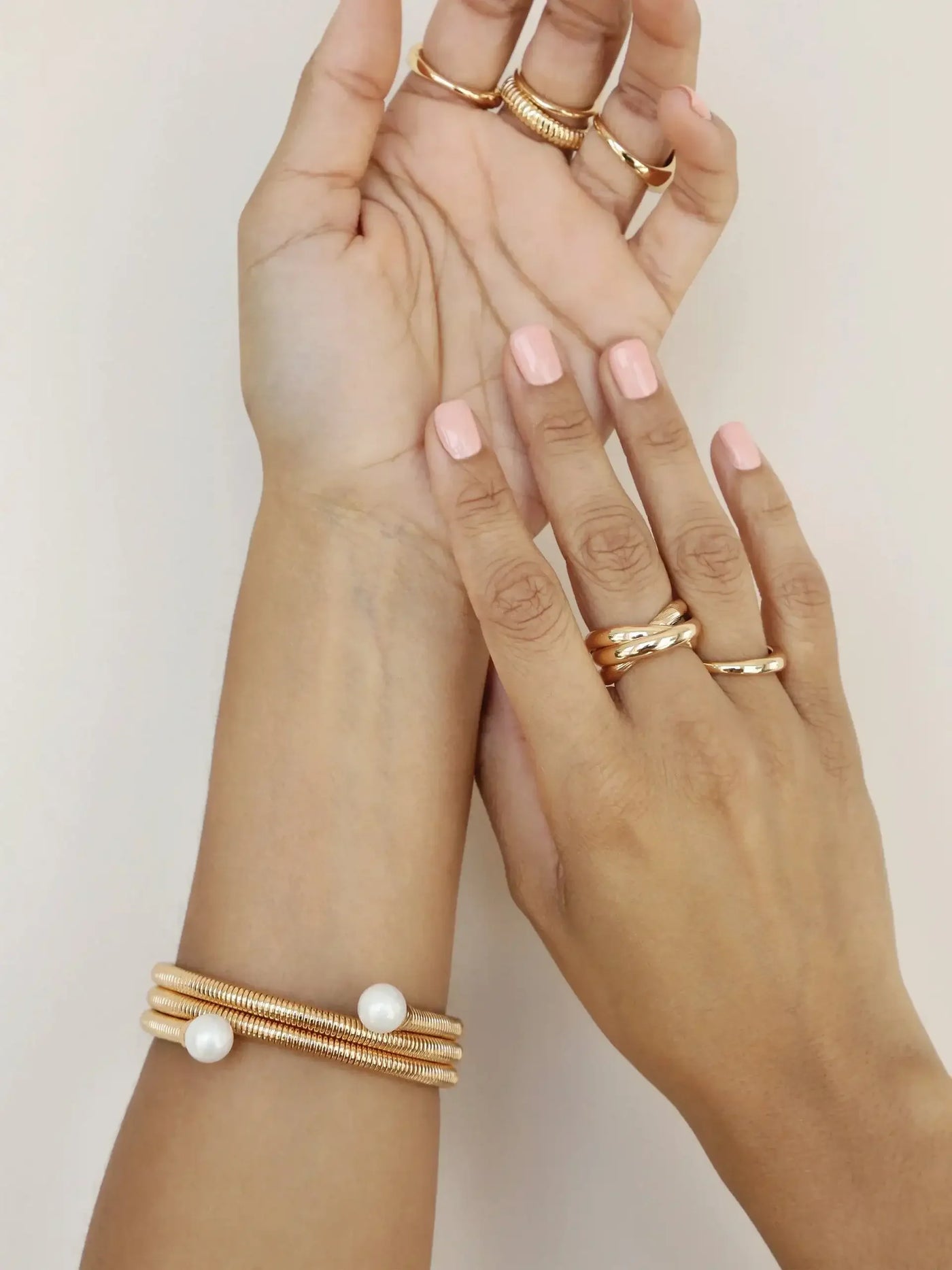 Two hands with manicured pink nails are adorned with multiple gold rings and bracelets, featuring a design that incorporates coils and white pearls, against a light neutral background.