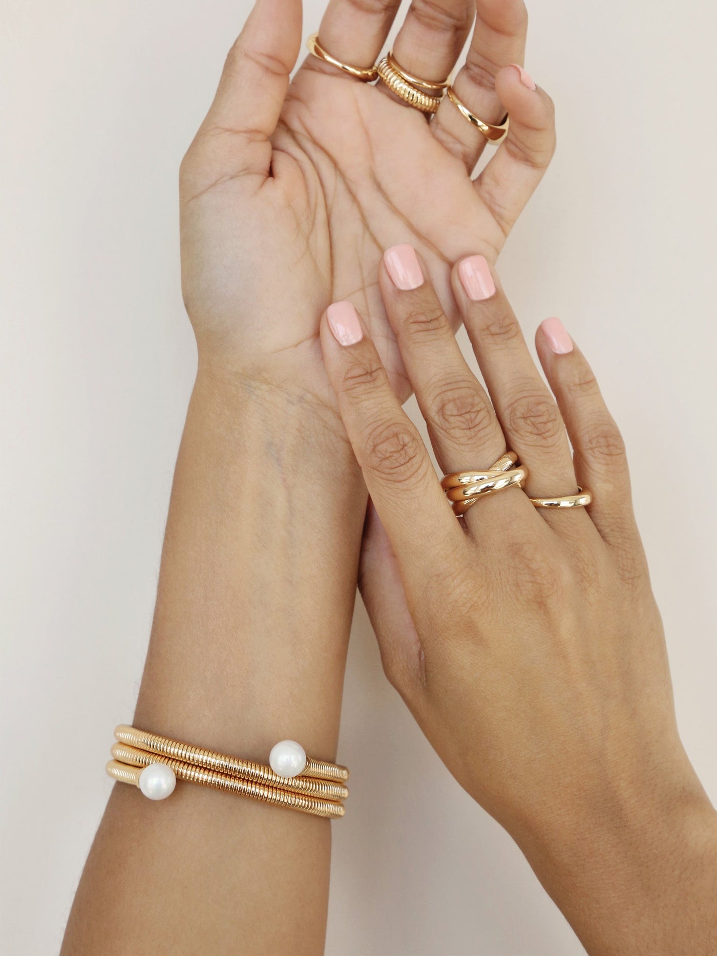 Two hands with manicured pink nails are adorned with multiple gold rings and bracelets, featuring a design that incorporates coils and white pearls, against a light neutral background.