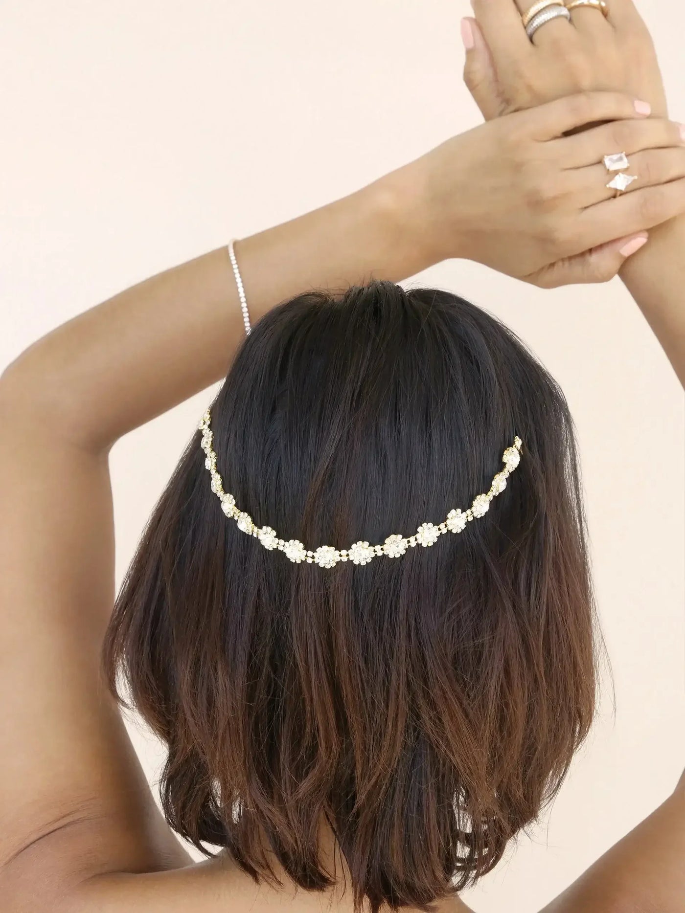 A woman with brown hair styles her hair by placing a decorative floral hairpiece at the back. Her hands, adorned with rings, are raised above her head. The background is a soft, neutral tone.