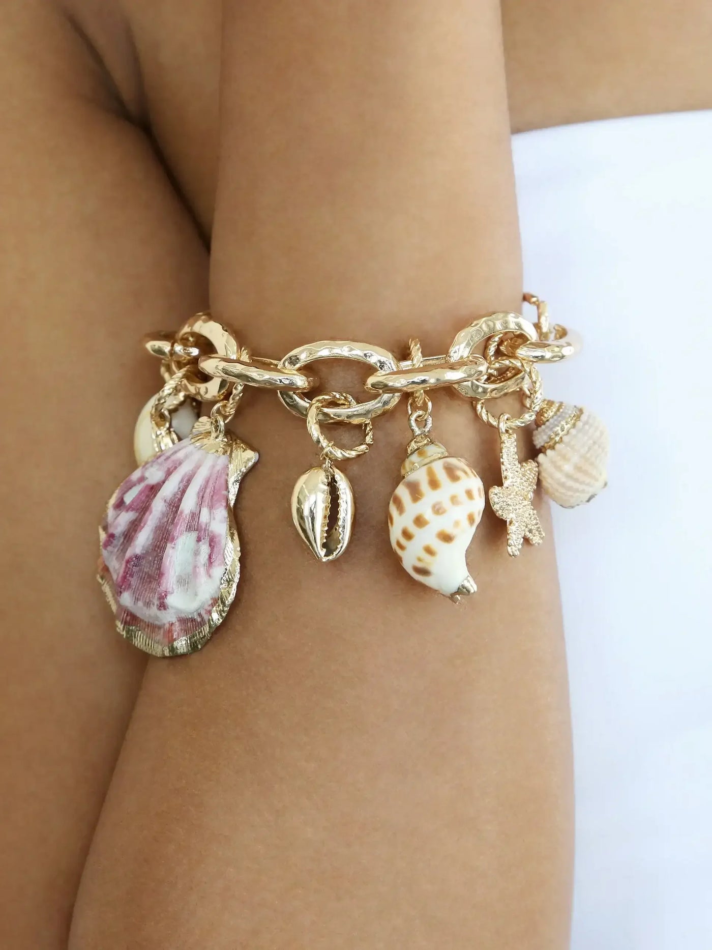 A gold chain bracelet features various decorative seashell charms, accentuating a tanned arm. The background is minimal, highlighting the bracelet's intricate details and beach-themed design.