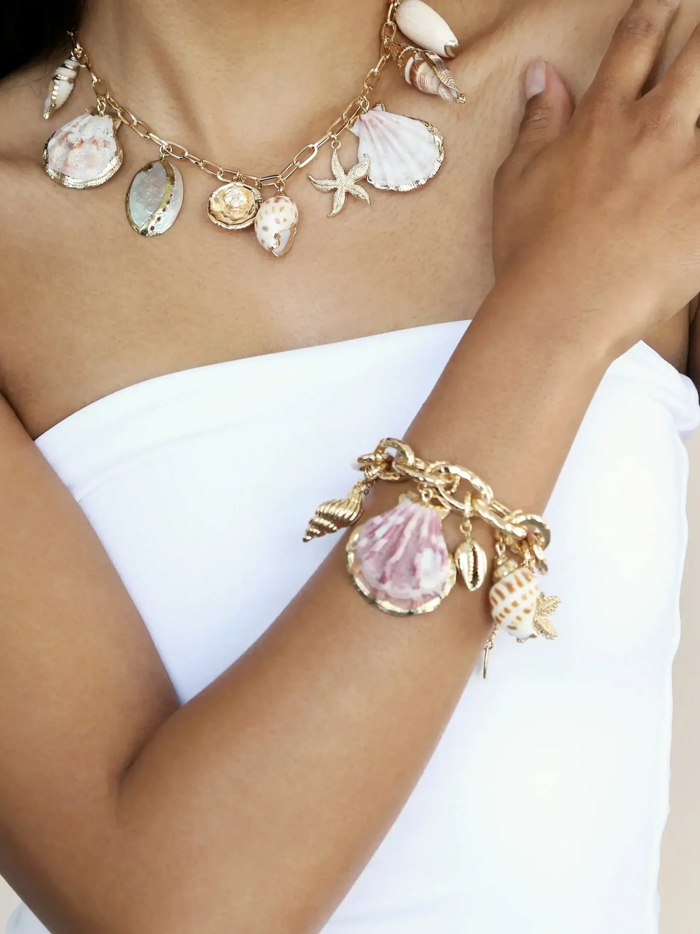 A necklace and bracelet adorned with various seashells and a starfish drape elegantly over a bare shoulder, creating a beach-like aesthetic against a neutral background.
