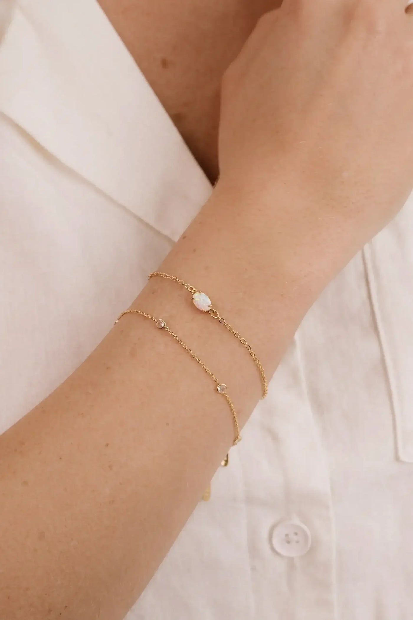 A hand with several delicate gold bracelets rests against a light-colored fabric, showcasing the jewelry’s shimmer against the skin, emphasizing elegance and simplicity in style.