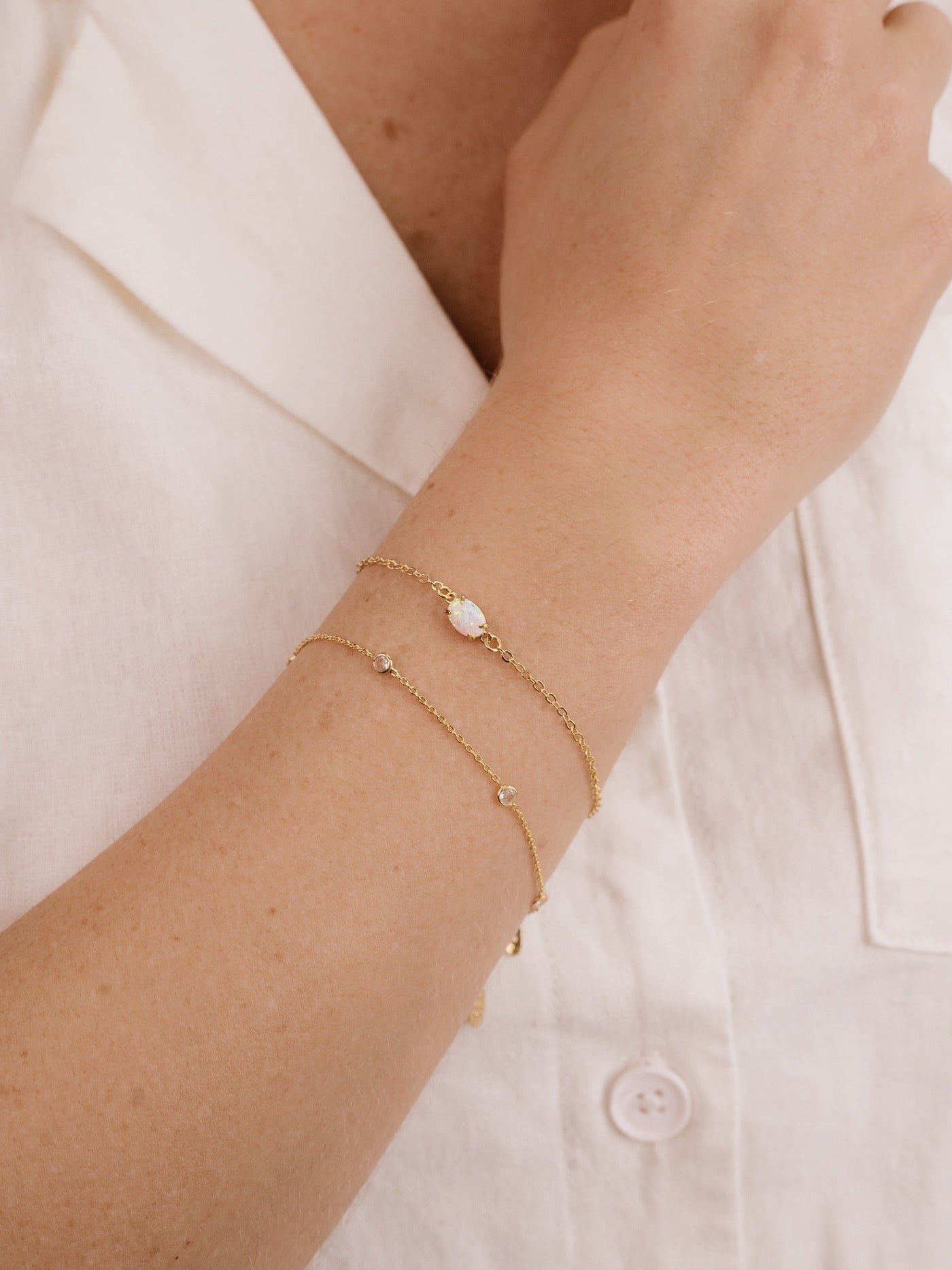 A hand with several delicate gold bracelets rests against a light-colored fabric, showcasing the jewelry’s shimmer against the skin, emphasizing elegance and simplicity in style.