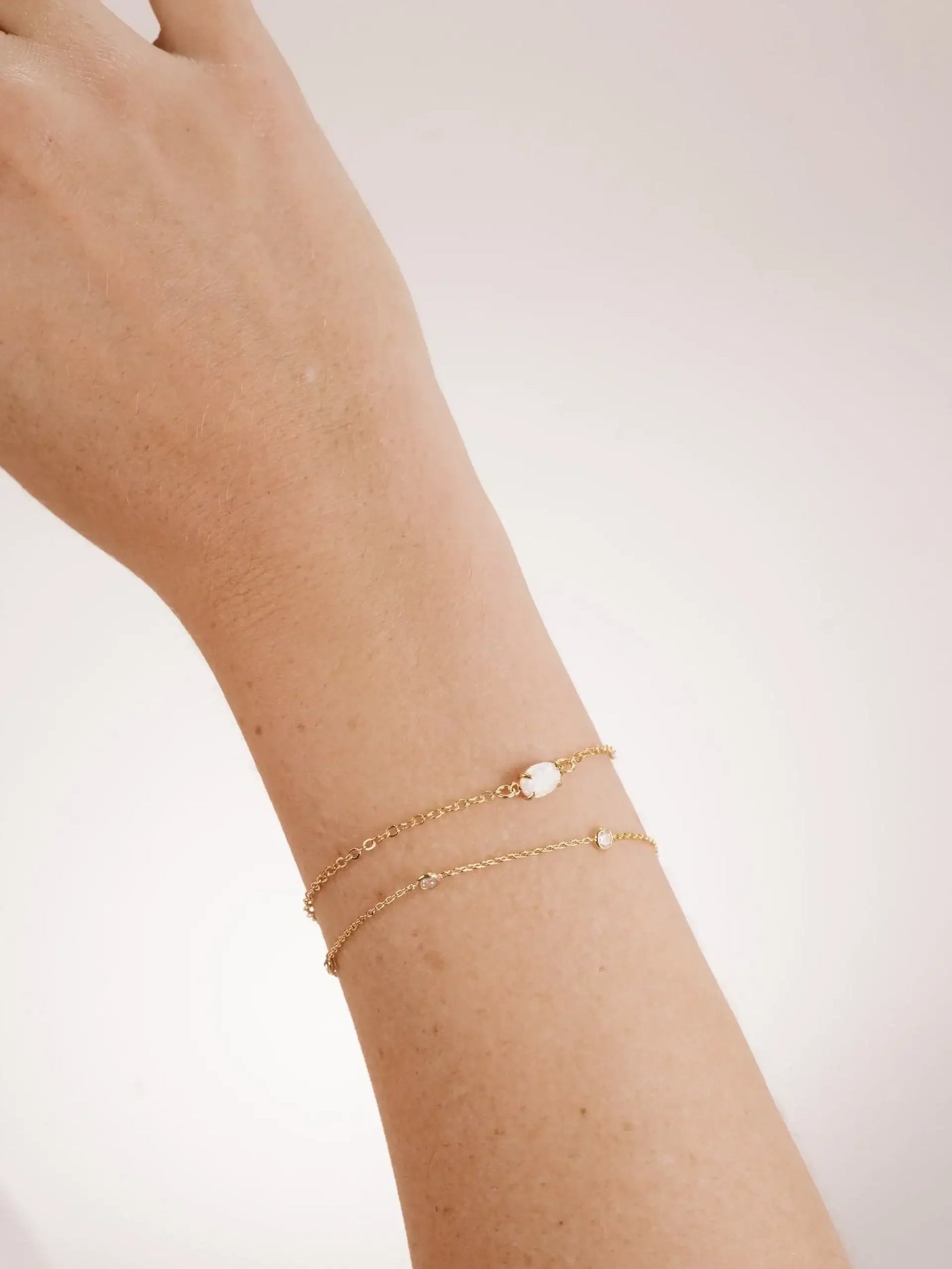 A hand with a gold chain bracelet is resting against a soft, light background. The bracelet features small, sparkling stones interspersed along the delicate chain.