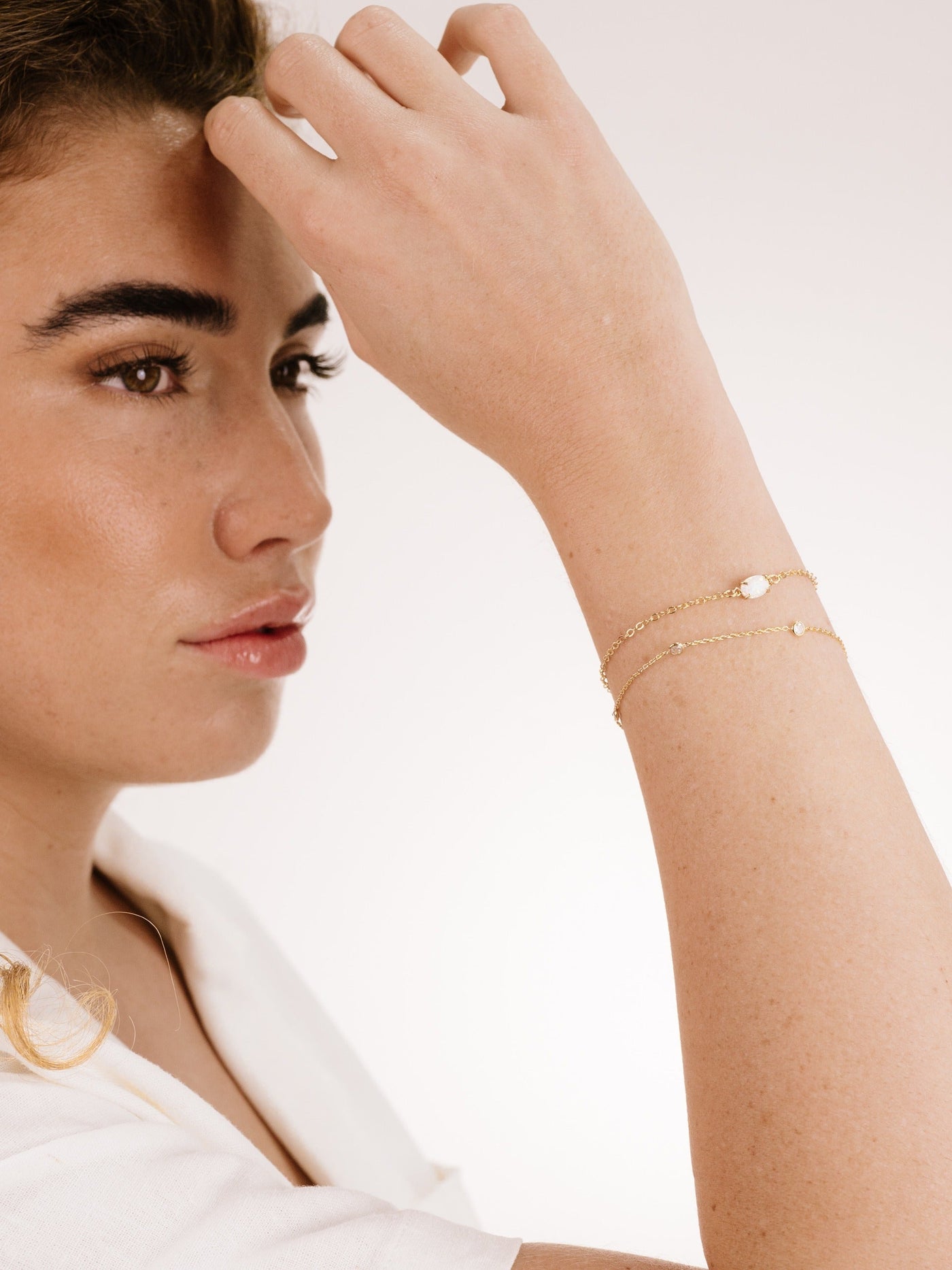 A woman with light brown hair is brushing it back with one hand, showcasing delicate gold bracelets on her wrist, against a softly lit, neutral background.
