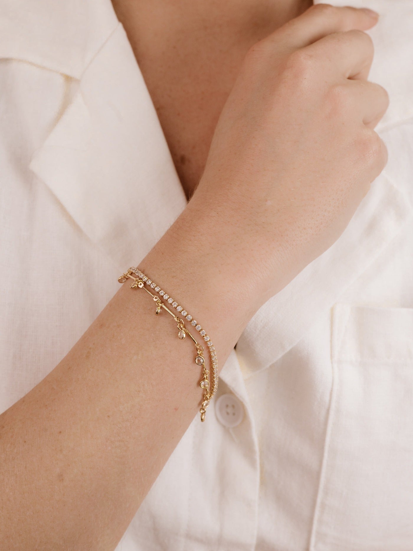 A gold bracelet adorned with small accents rests on a wrist, enhancing the arm's elegance. The wearer is dressed in a light-colored, buttoned shirt, suggesting a casual yet refined setting.