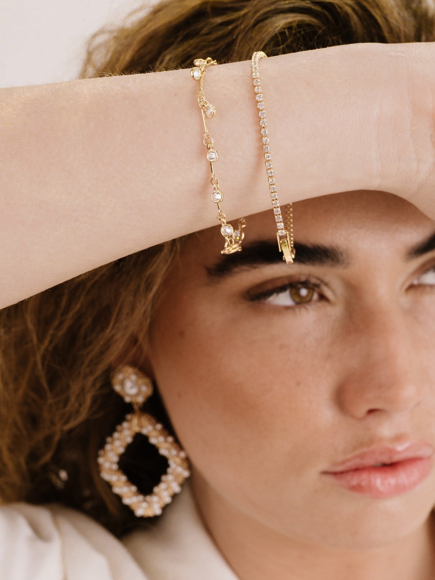 A hand adorned with two elegant gold bracelets is raised near a woman's face, showcasing luxurious jewelry, while she gazes thoughtfully. A large pearl earring complements her look.