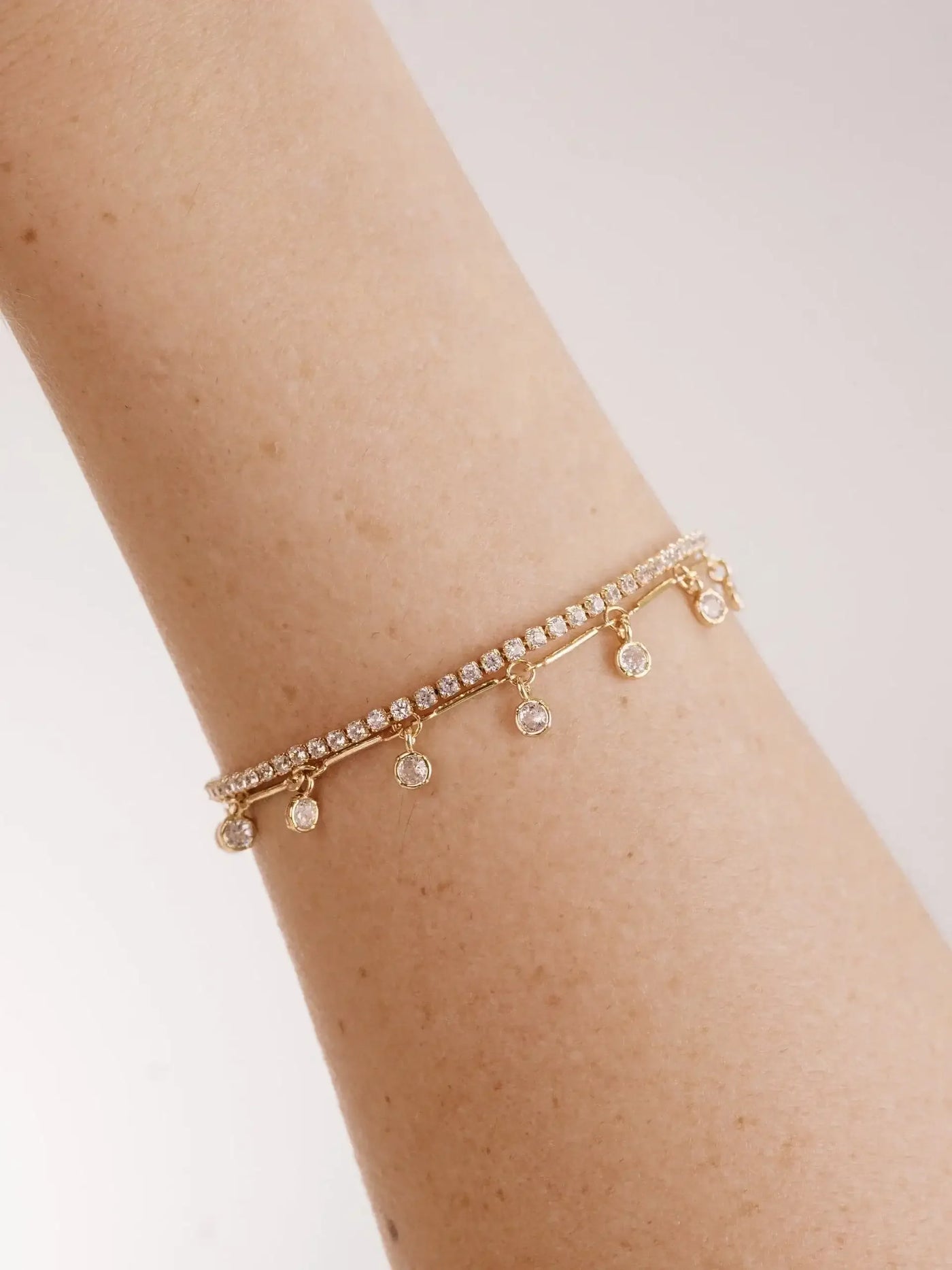 A gold bracelet adorned with small, sparkling stones hangs delicately on a light-skinned arm. The bracelet features dangling gems, reflecting light against a soft, neutral background.