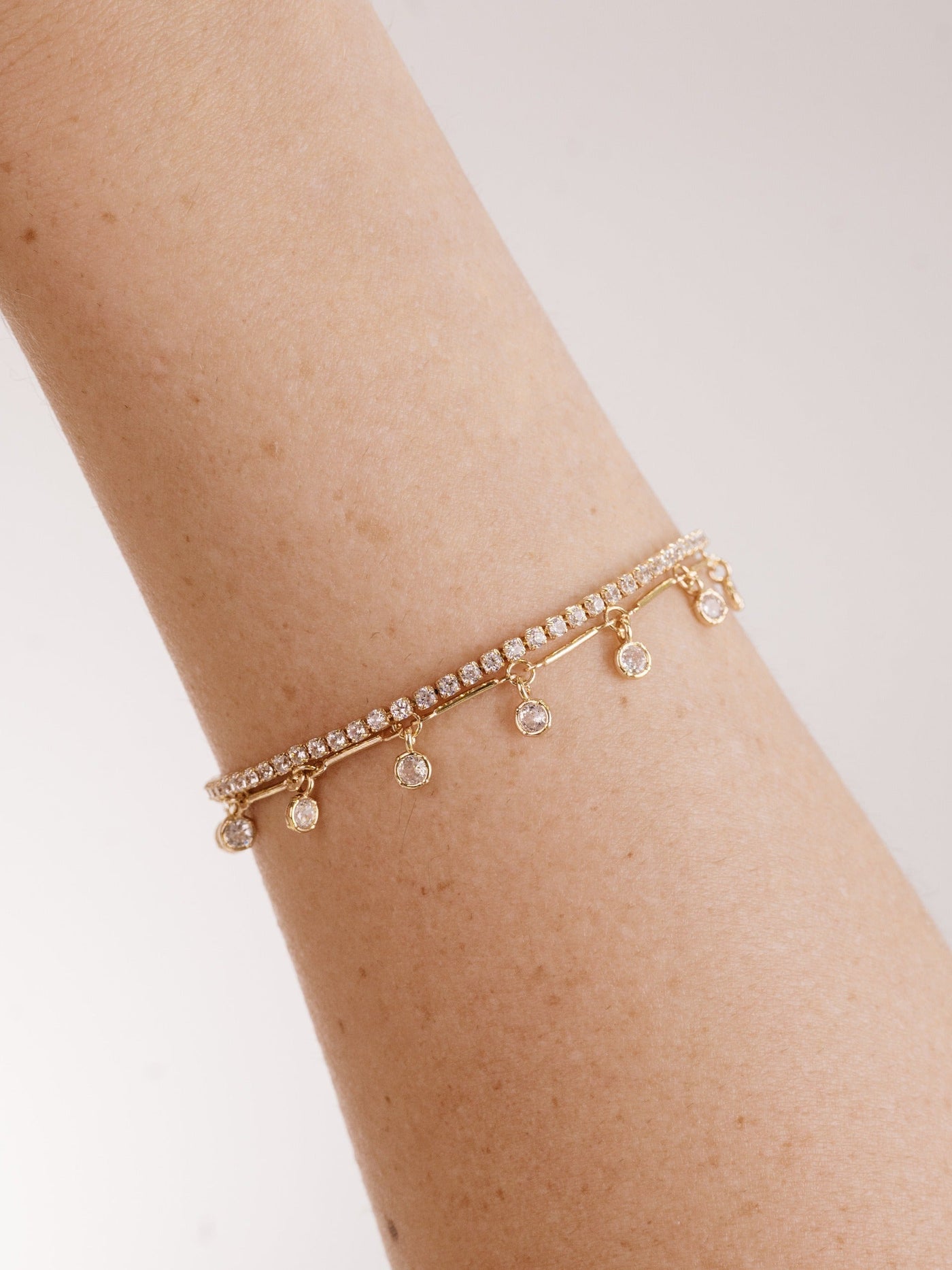 A gold bracelet adorned with small, sparkling stones hangs delicately on a light-skinned arm. The bracelet features dangling gems, reflecting light against a soft, neutral background.