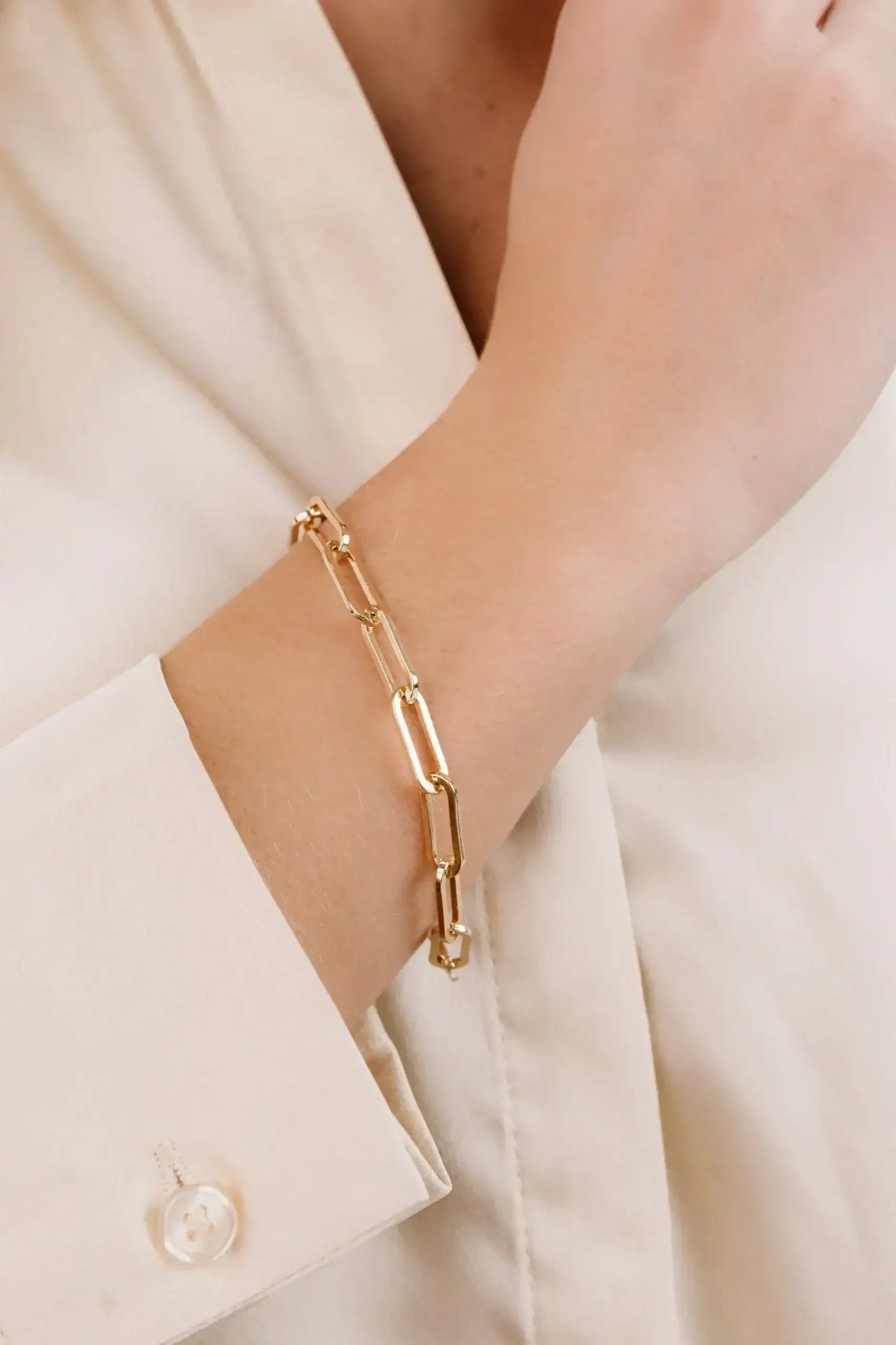 A gold chain bracelet adorns a wrist, highlighting its elegant links. The arm, partially covered by a light-colored blouse, rests against a neutral backdrop.