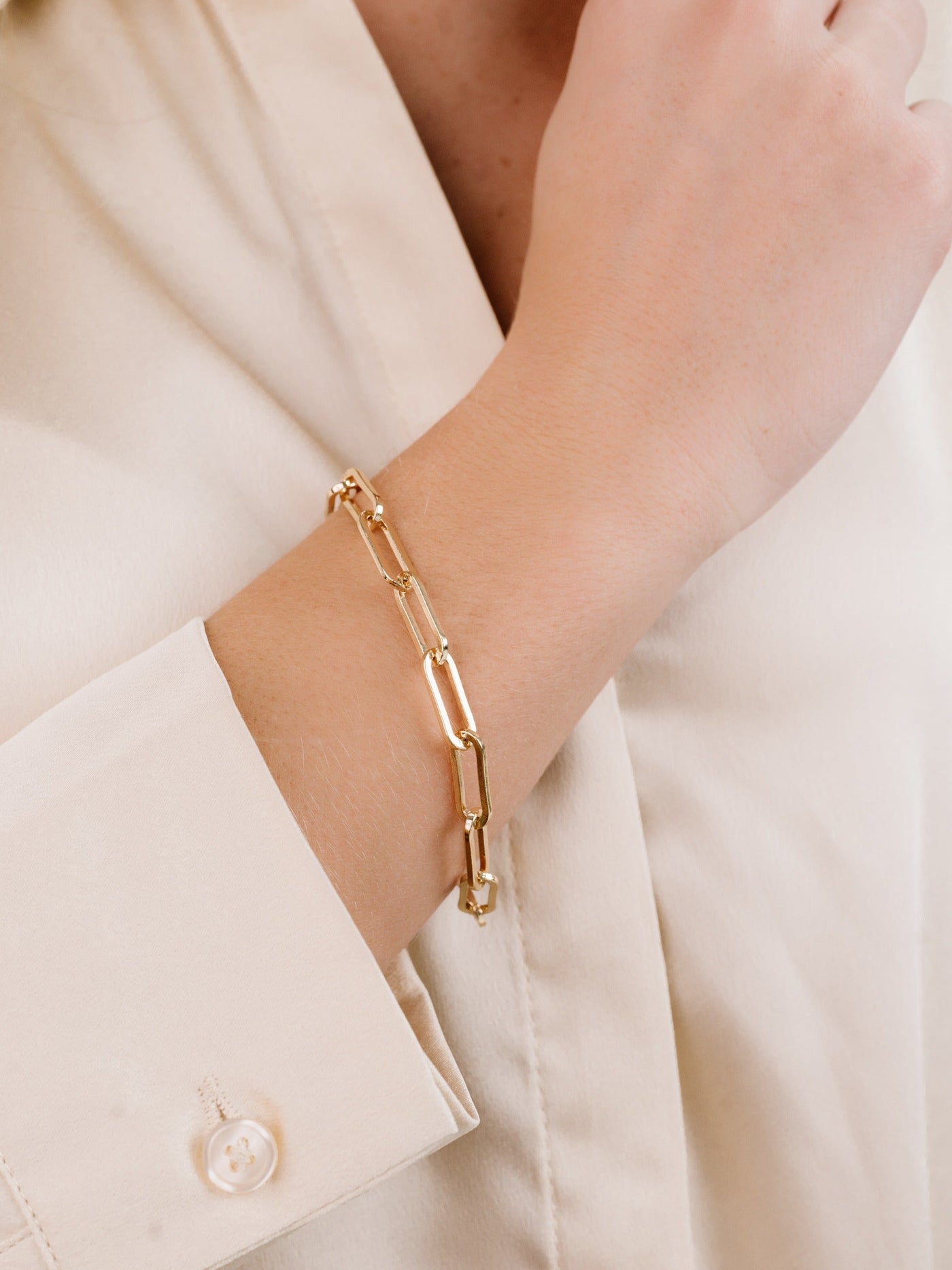 A gold chain bracelet adorns a wrist, highlighting its elegant links. The arm, partially covered by a light-colored blouse, rests against a neutral backdrop.