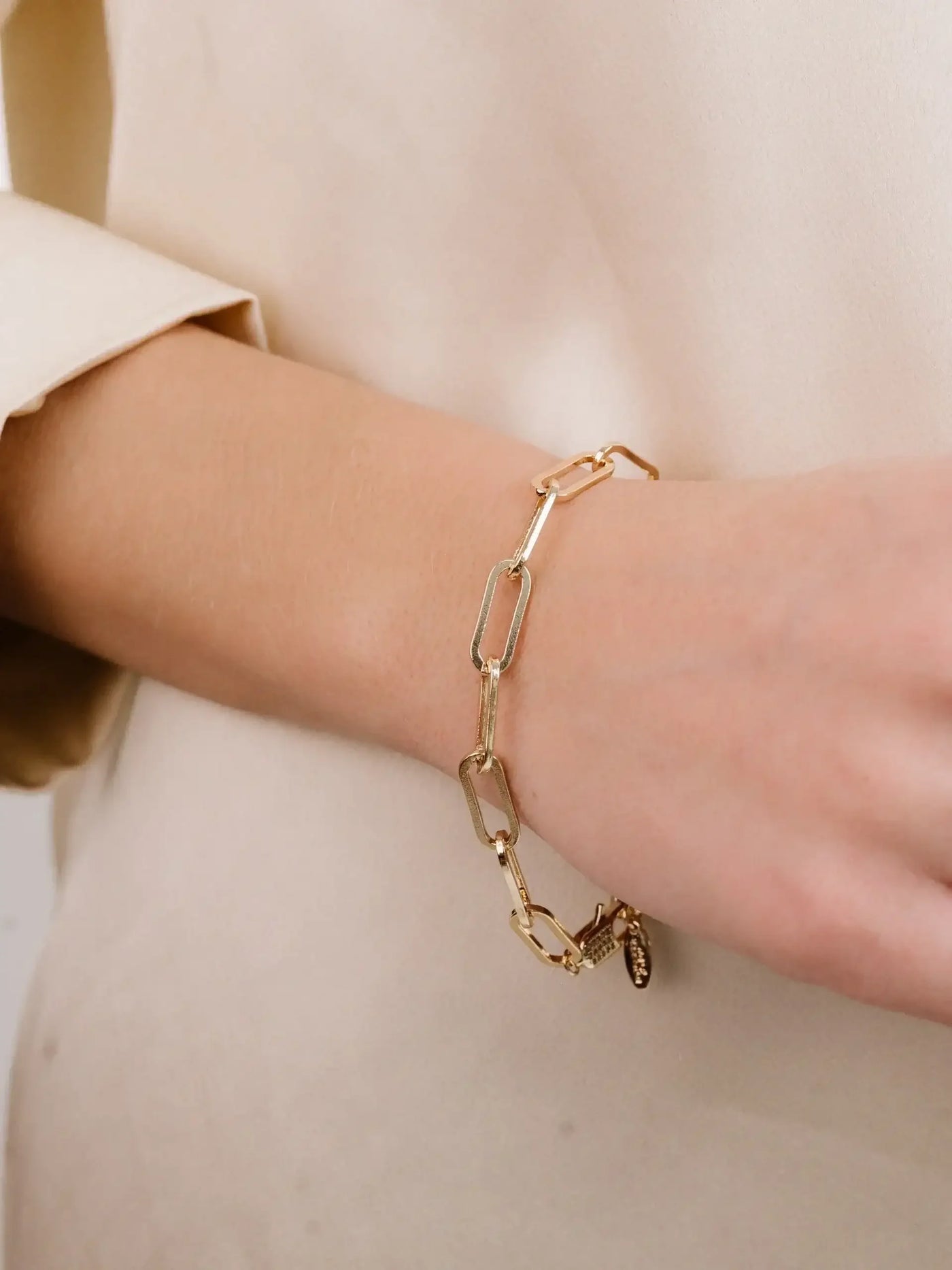 A gold link bracelet adorns a person's wrist while their hand rests against a soft, neutral-colored fabric. The setting appears elegant and understated.