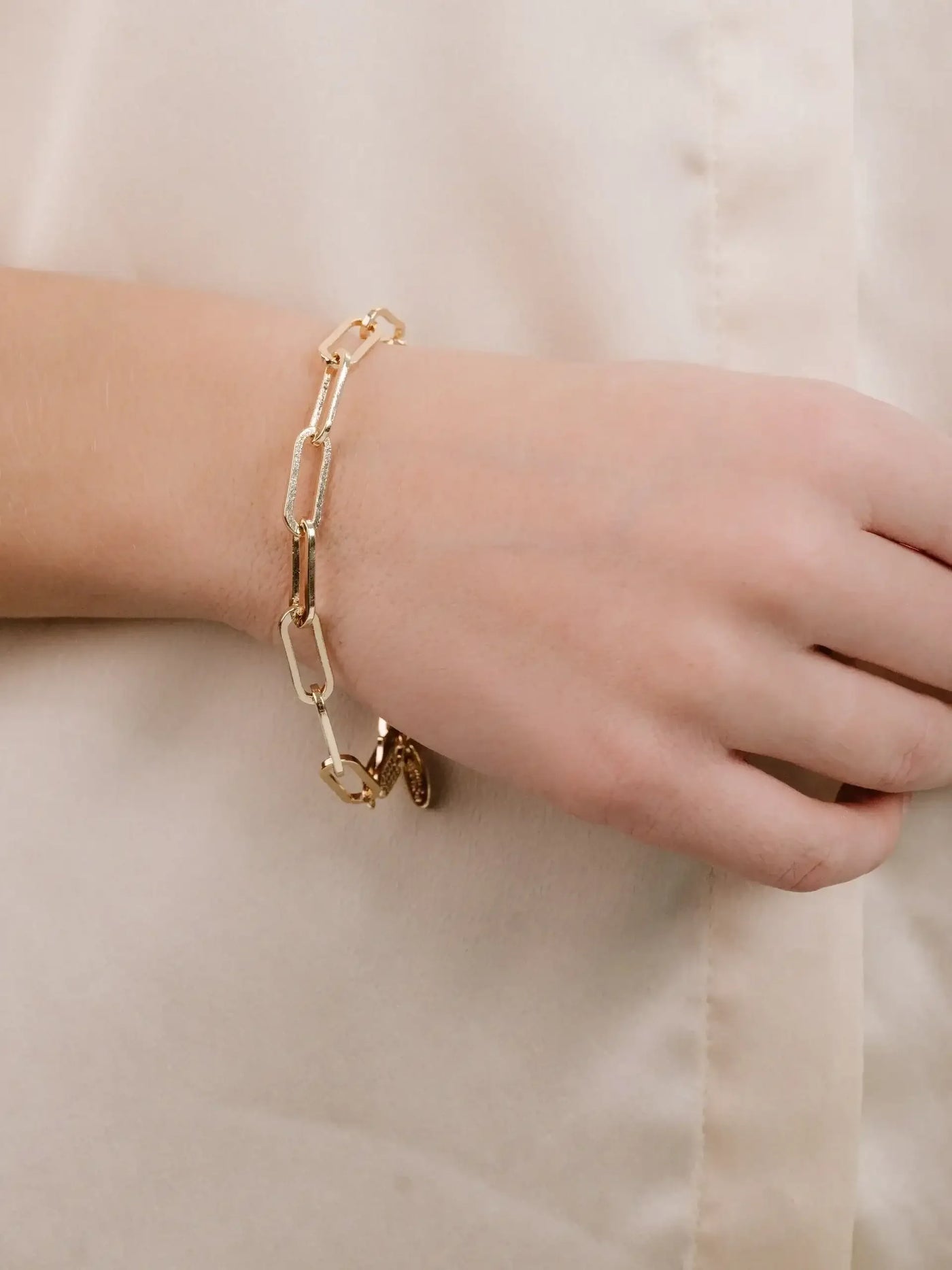 A gold link bracelet adorns a wrist as the hand rests against a soft, neutral-colored fabric, suggesting elegance and style in a simple, minimalist setting.