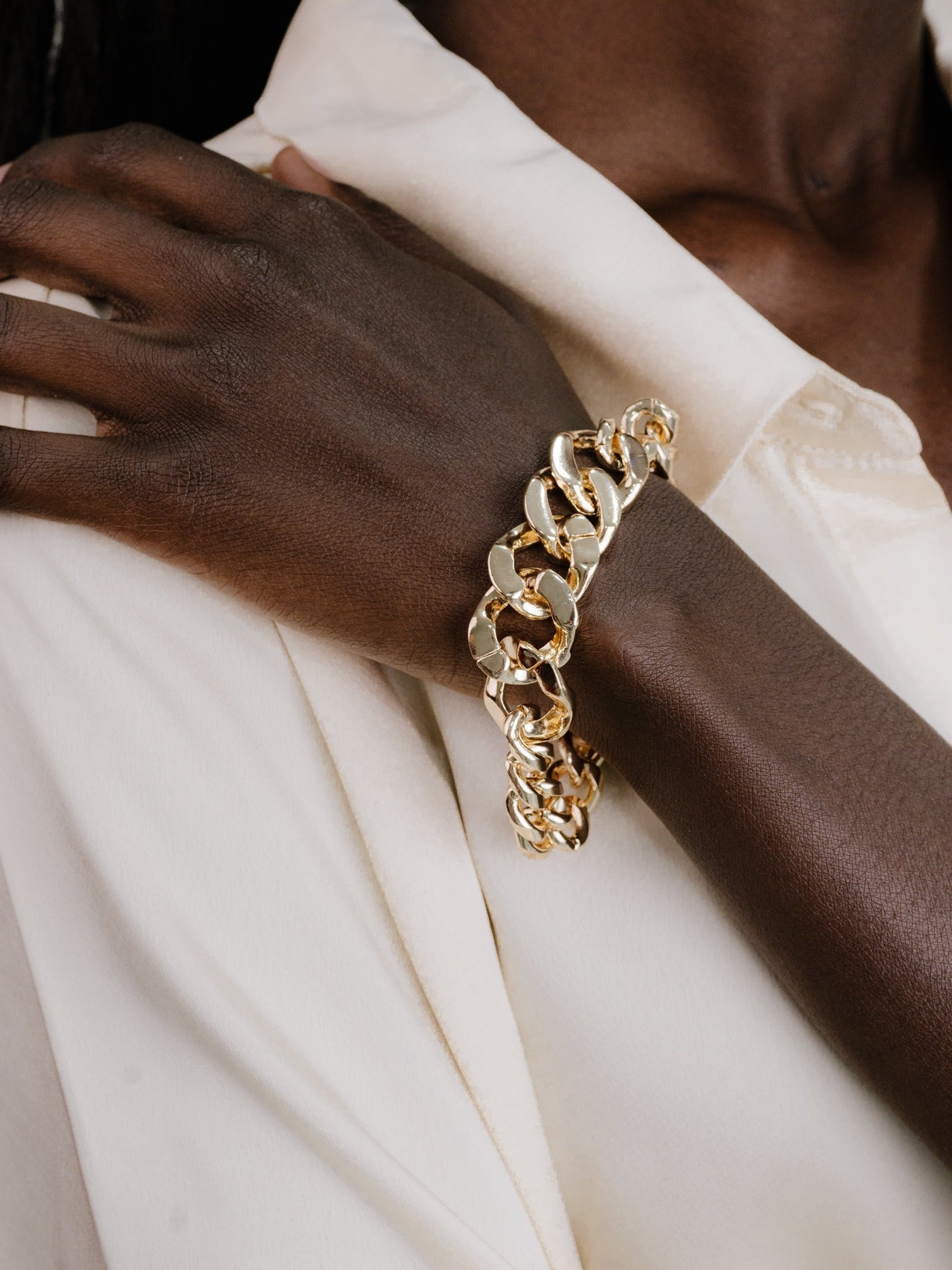 A gold chain bracelet adorns a dark-skinned hand resting gently on a cream-colored shirt, showcasing the contrasting textures and colors of the jewelry and fabric.