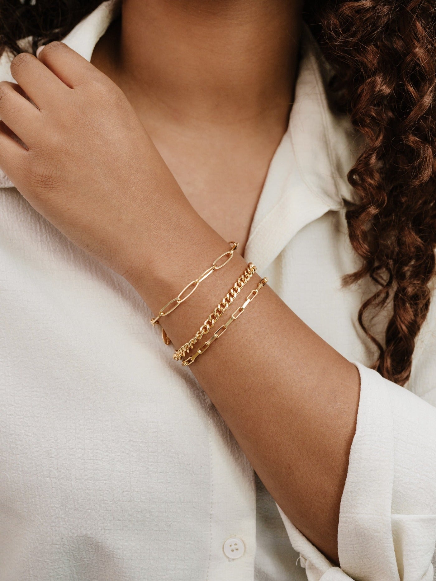 A hand adorned with three gold bracelets rests against a textured white shirt. The bracelets vary in design, reflecting light and showcasing a stylish complement to the outfit.