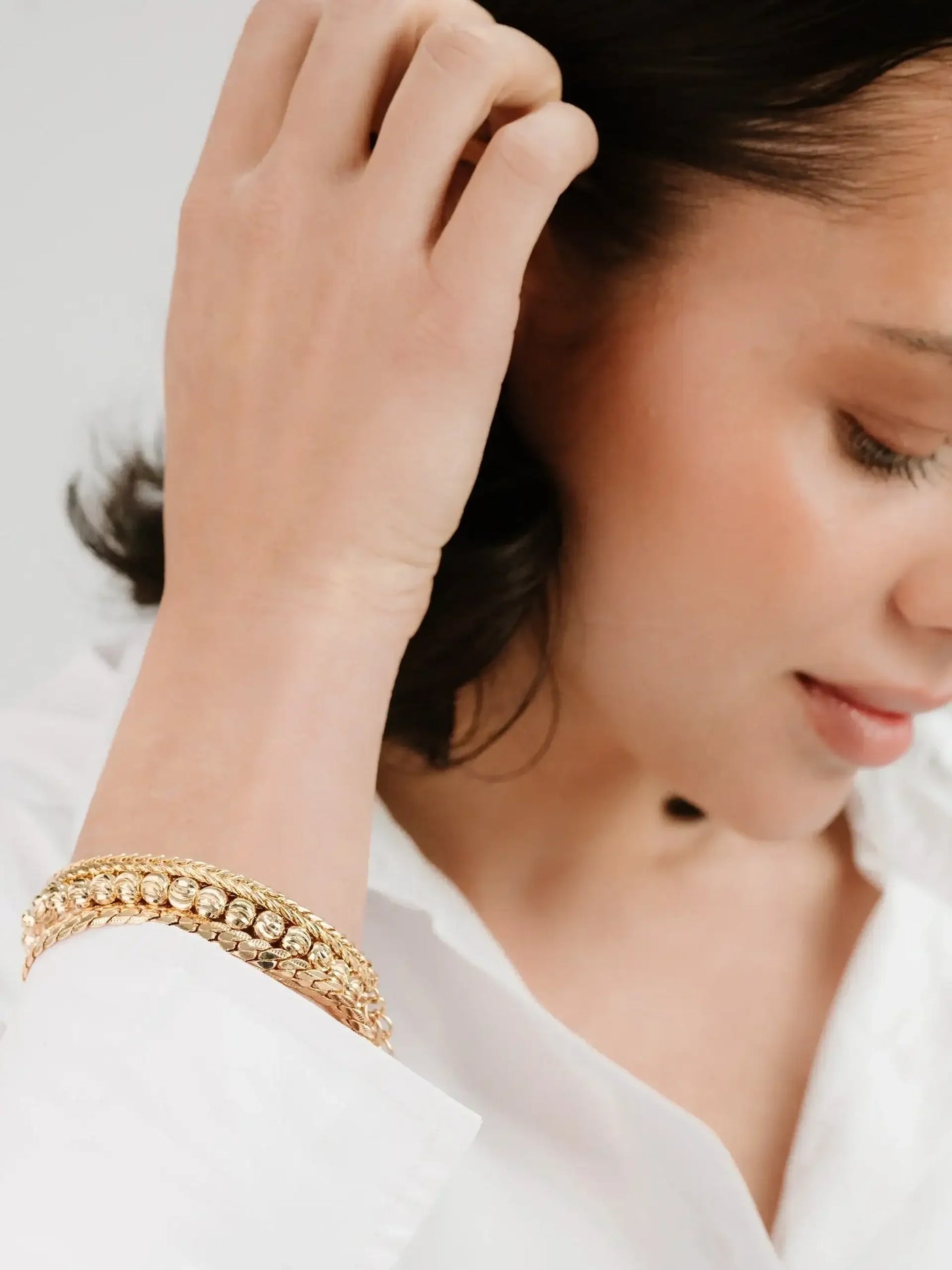 A woman with a refined appearance is adjusting her hair with one hand while wearing multiple gold bracelets on her wrist, dressed in a white shirt, against a subtle, light background.