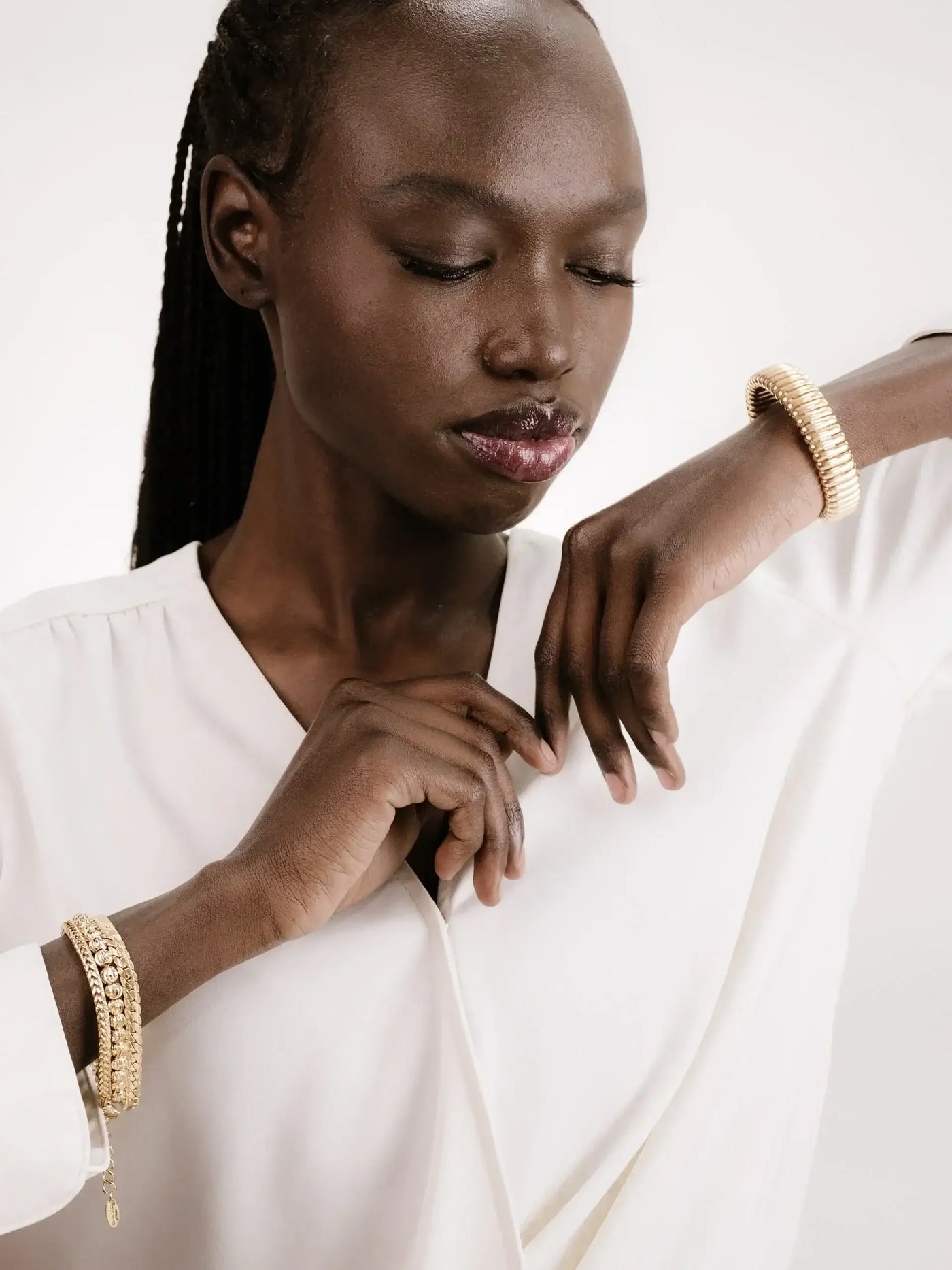 A person with long black hair wears a white shirt and gold bracelets, gently touching their collar while gazing thoughtfully downwards against a light, neutral background.