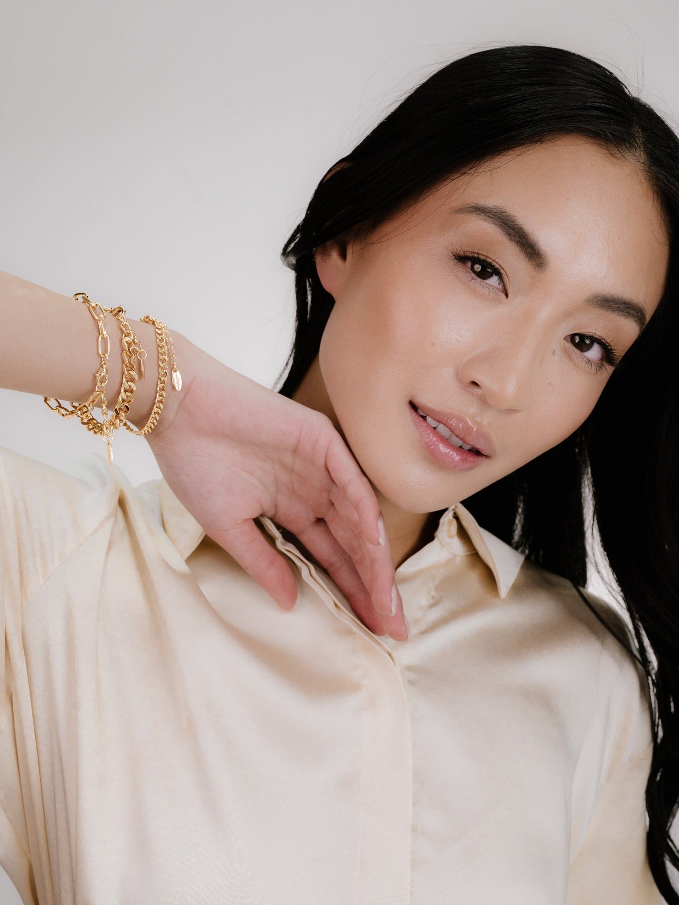 A woman with long black hair wears a light-colored shirt, showcasing multiple gold bracelets on her wrist while gently touching her face, set against a soft, neutral background.