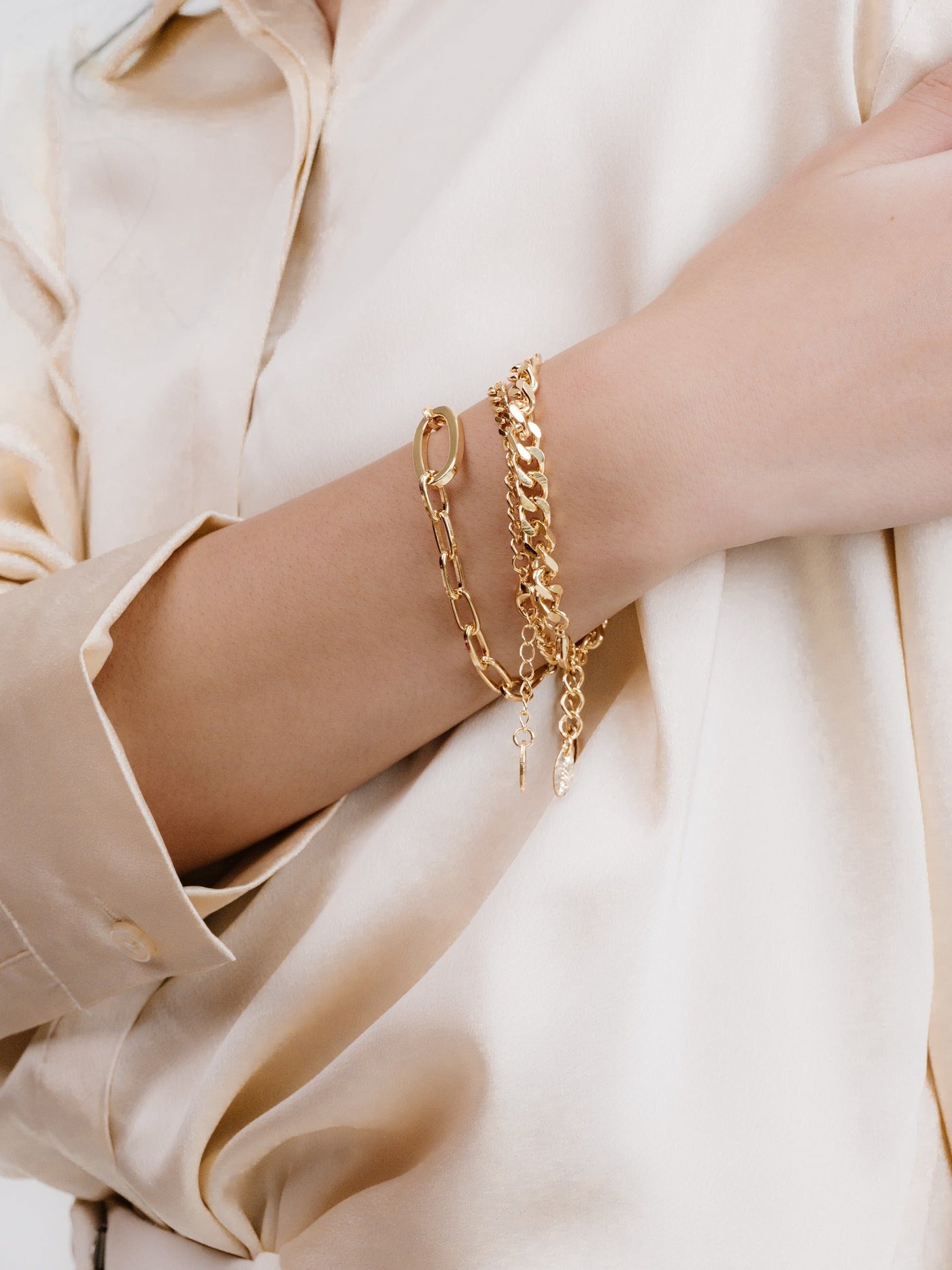 A hand adorned with multiple gold bracelets rests against a light, silky fabric. The bracelets feature varying chains and designs, showcasing a blend of elegance and style.