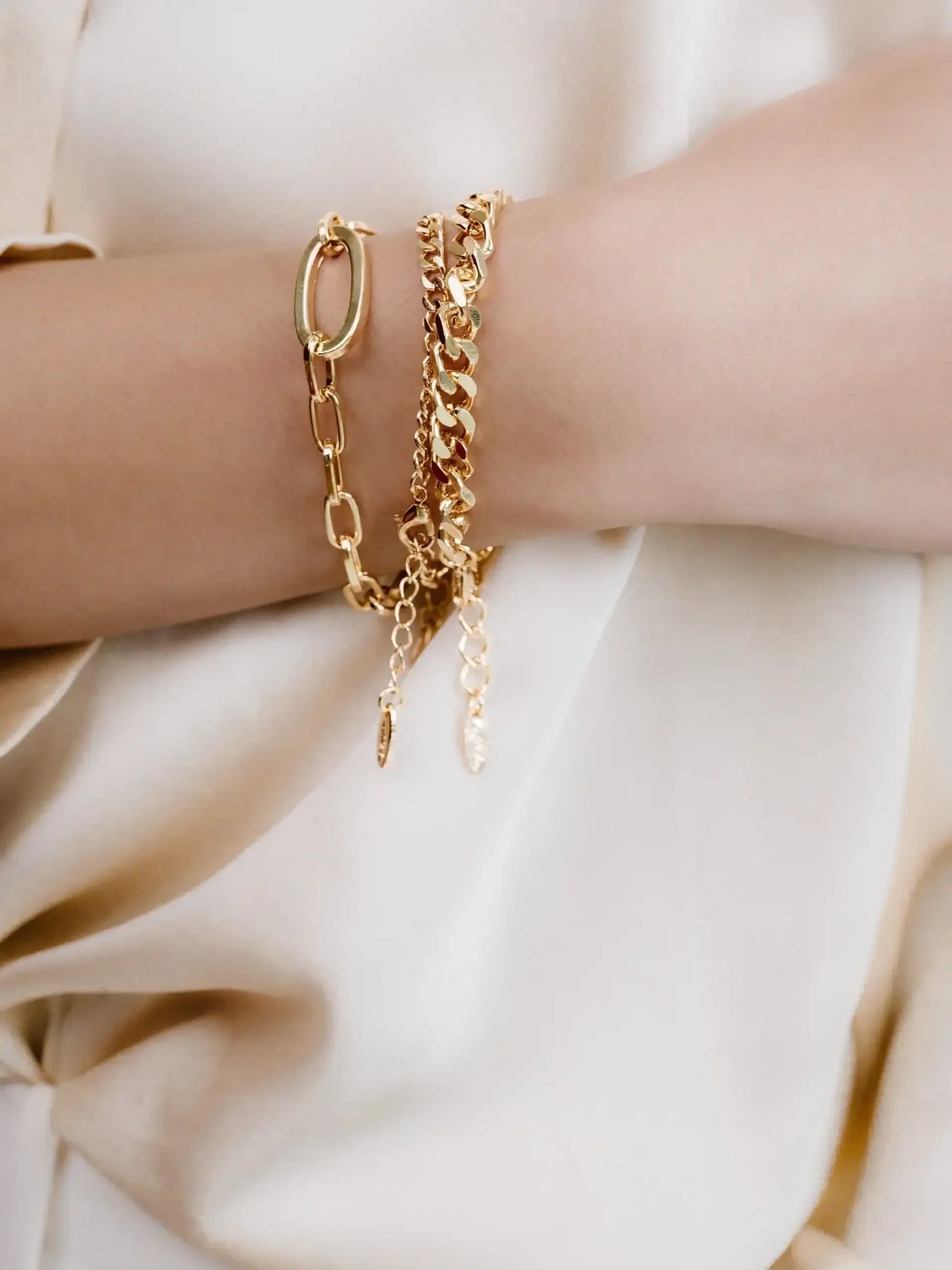 A hand displays several gold bracelets, including a chain and a delicate design with pendants, resting against a cream-colored fabric, highlighting the jewelry's shine and elegance.