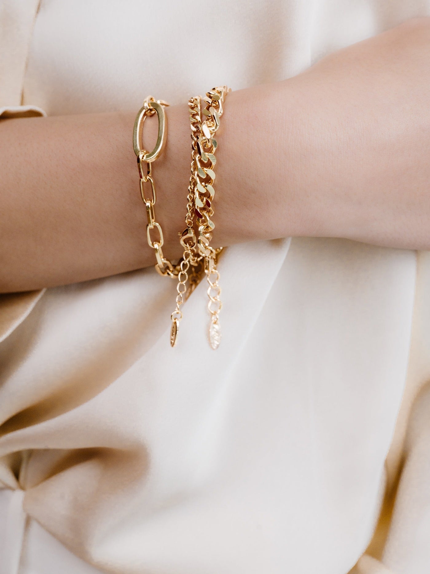 A hand displays several gold bracelets, including a chain and a delicate design with pendants, resting against a cream-colored fabric, highlighting the jewelry's shine and elegance.