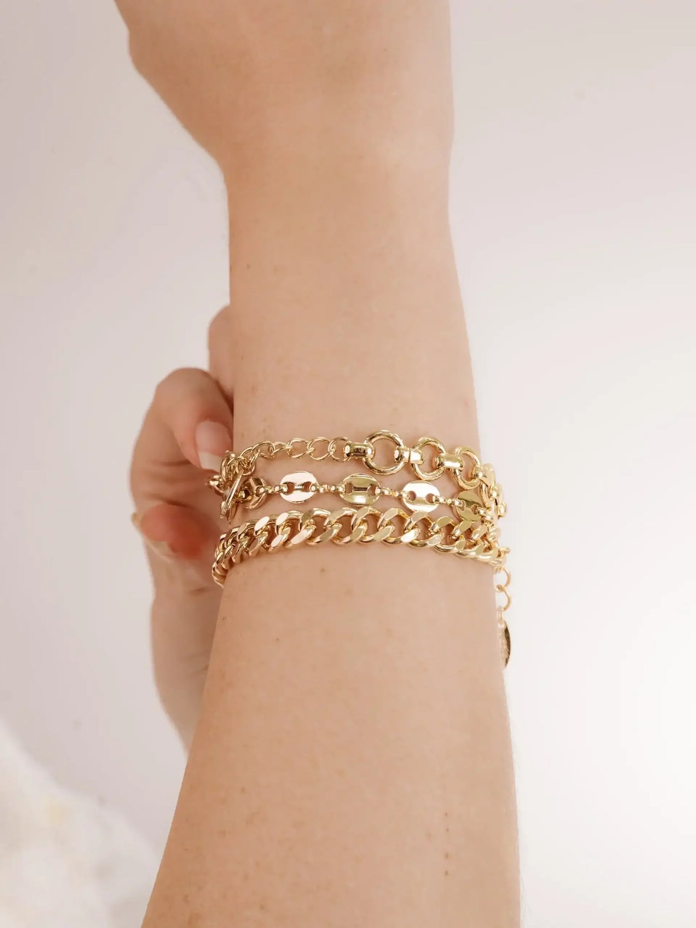 Gold bracelets adorn a wrist, with multiple chains layered together. The hand is positioned slightly raised, in a soft focus against a light background, emphasizing the jewelry's shine.