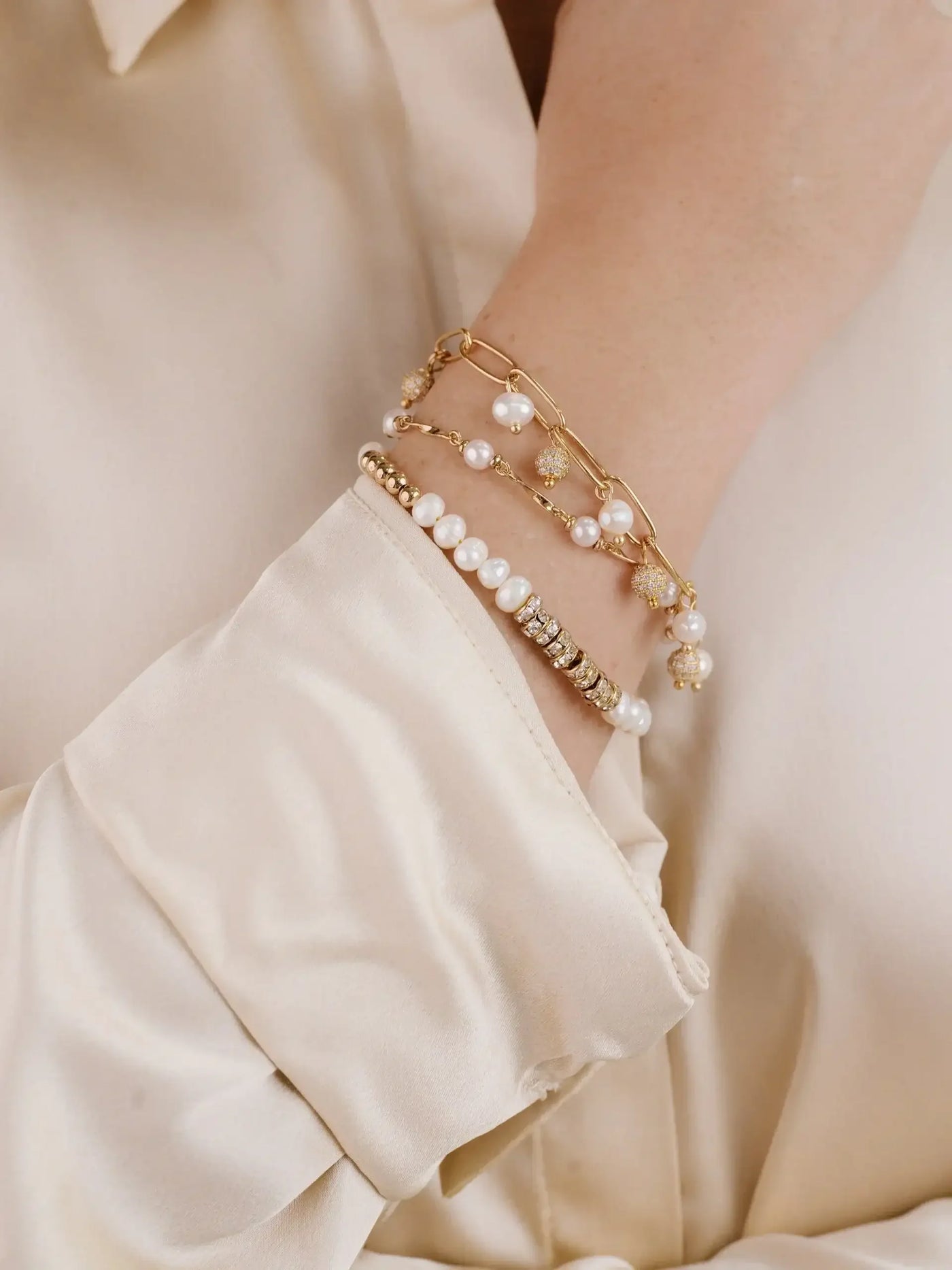 A hand adorned with multiple elegant bracelets, including pearls and gold accents, rests against a softly draped, cream-colored fabric. The arm's gentle posture contrasts with the smoothness of the material.