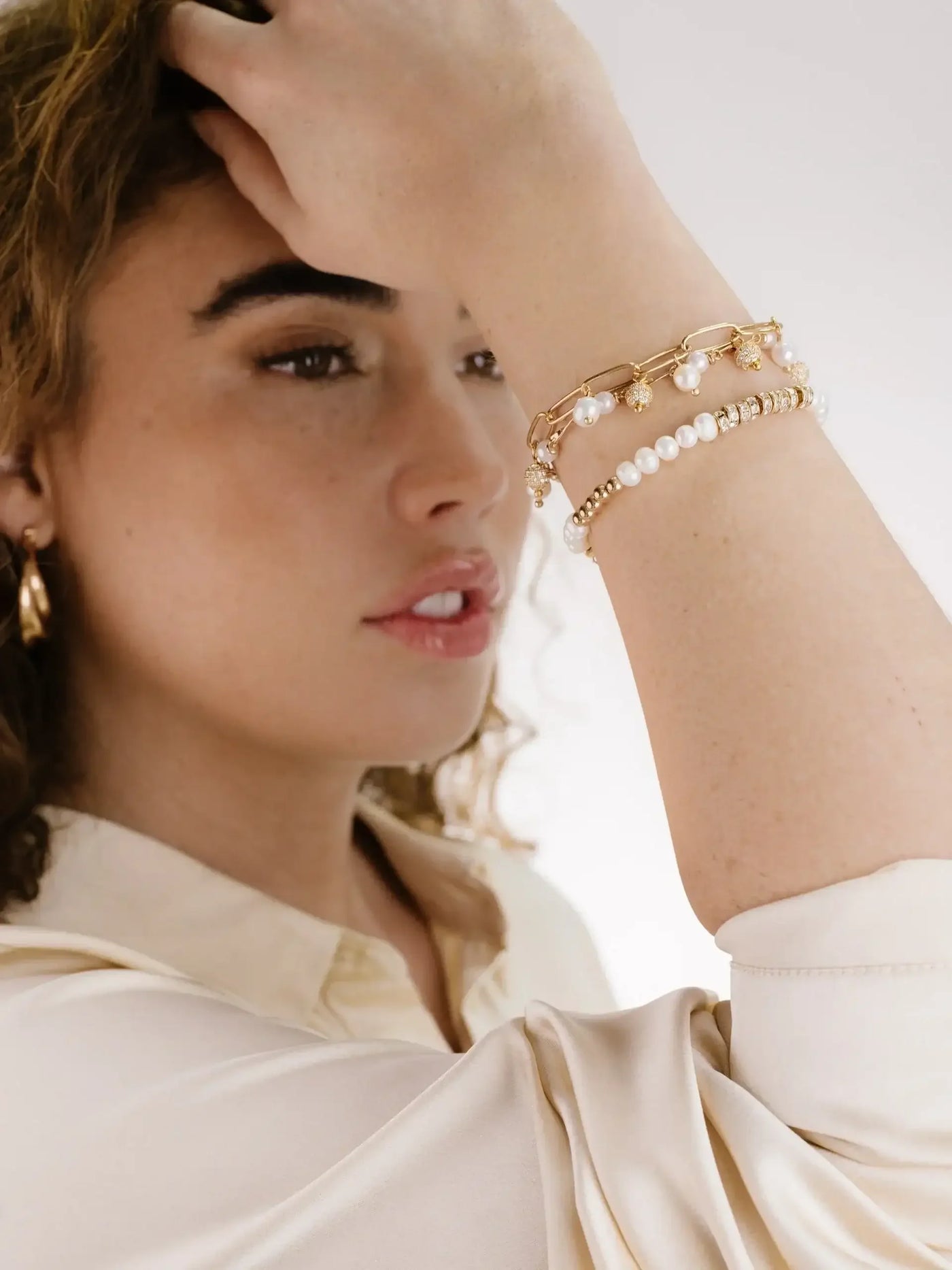 A woman with curly hair touches her forehead, showcasing multiple bracelets on her wrist. She is dressed in a light-colored blouse, set against a softly blurred background.