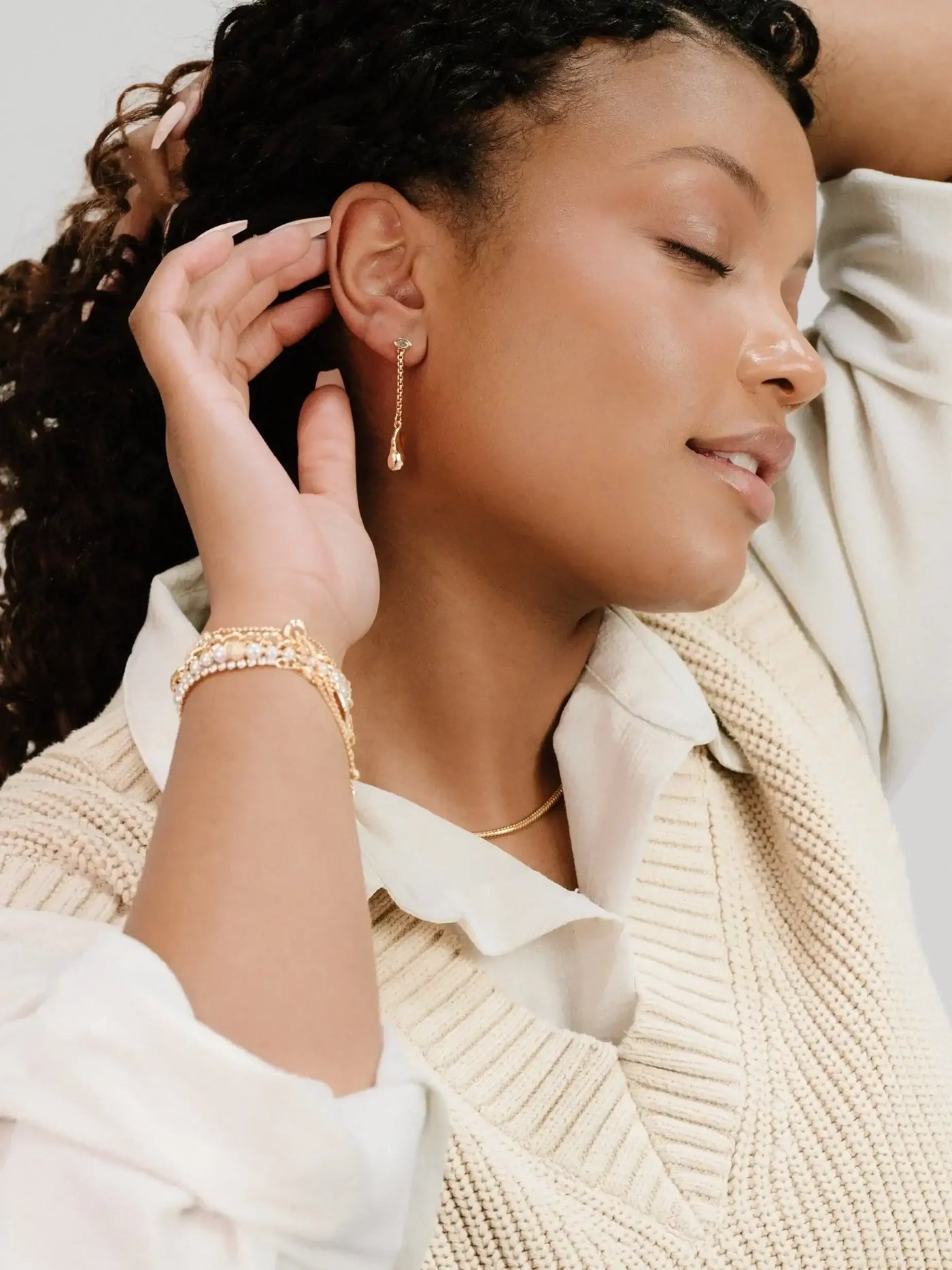A woman with curly hair delicately touches her ear, showcasing a dangling earring. She wears layered bracelets and a textured knit sweater, set against a soft, neutral background.