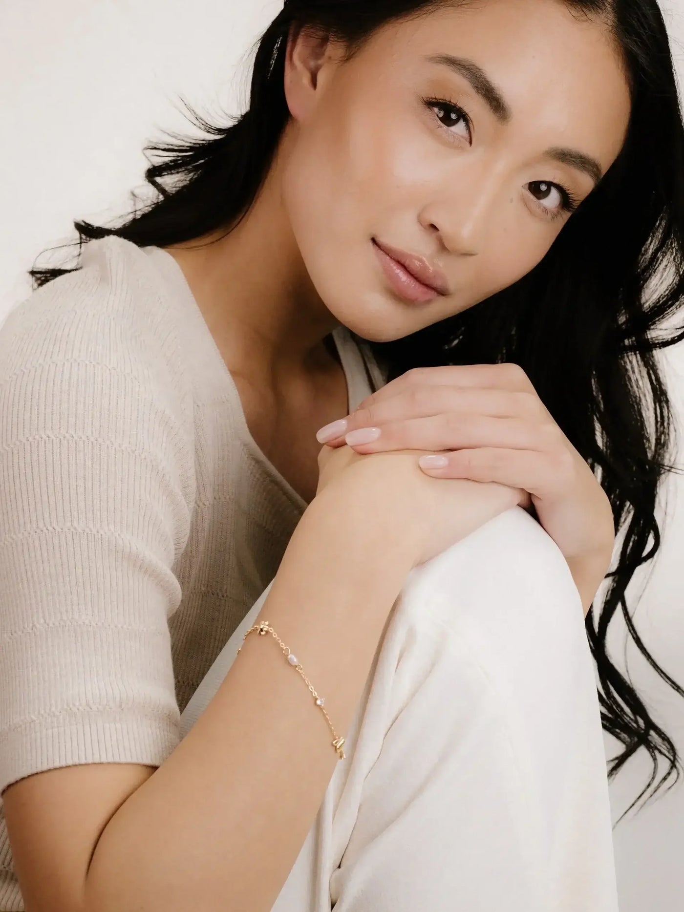 A young woman with long, dark hair rests her chin on her knees, wearing a light, textured top. She flaunts a delicate gold bracelet against a soft, neutral background.