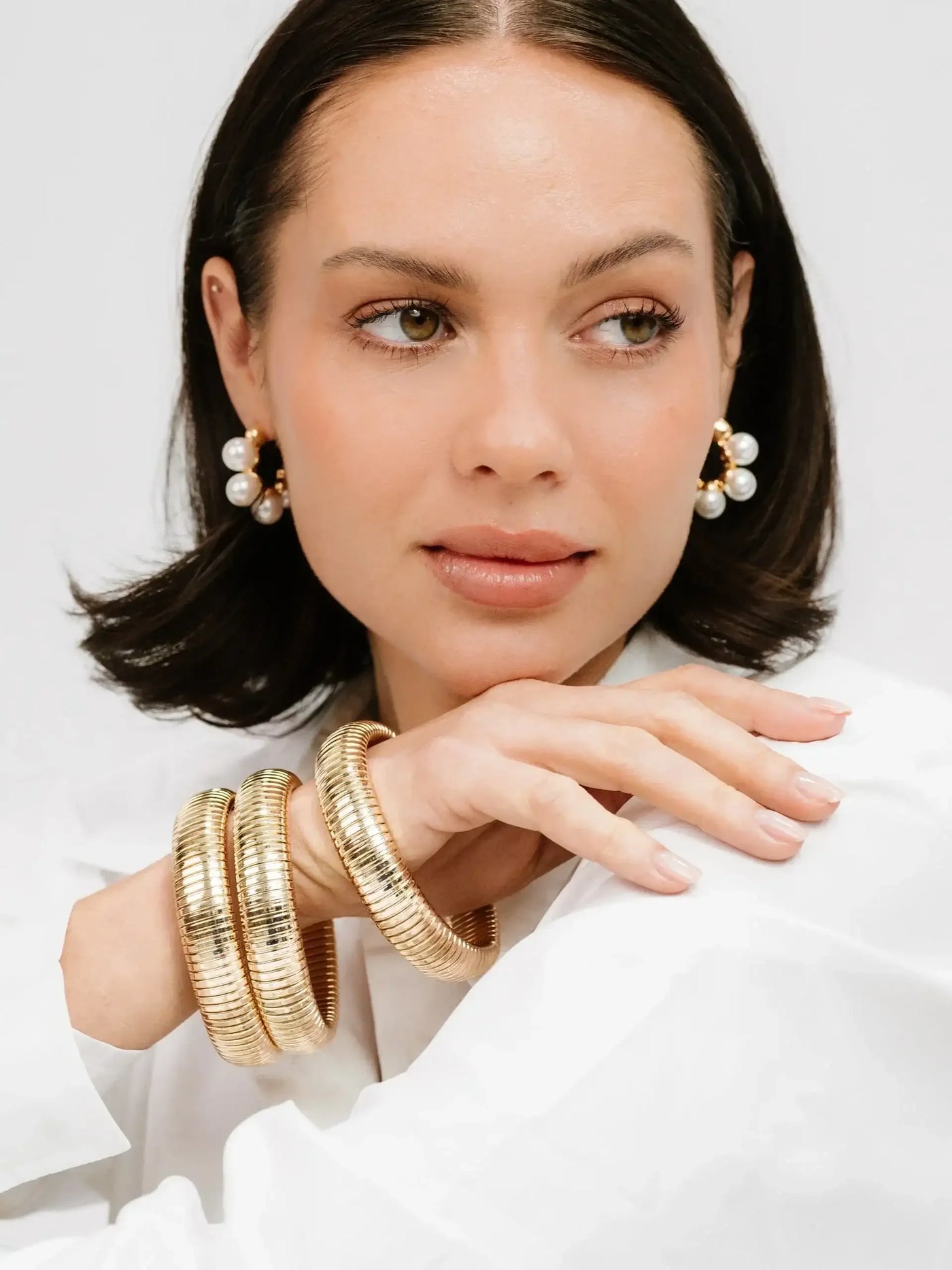 A woman with shoulder-length dark hair poses softly, wearing large pearl earrings and three gold bangles, with her hand resting elegantly on a white fabric. The background is plain.