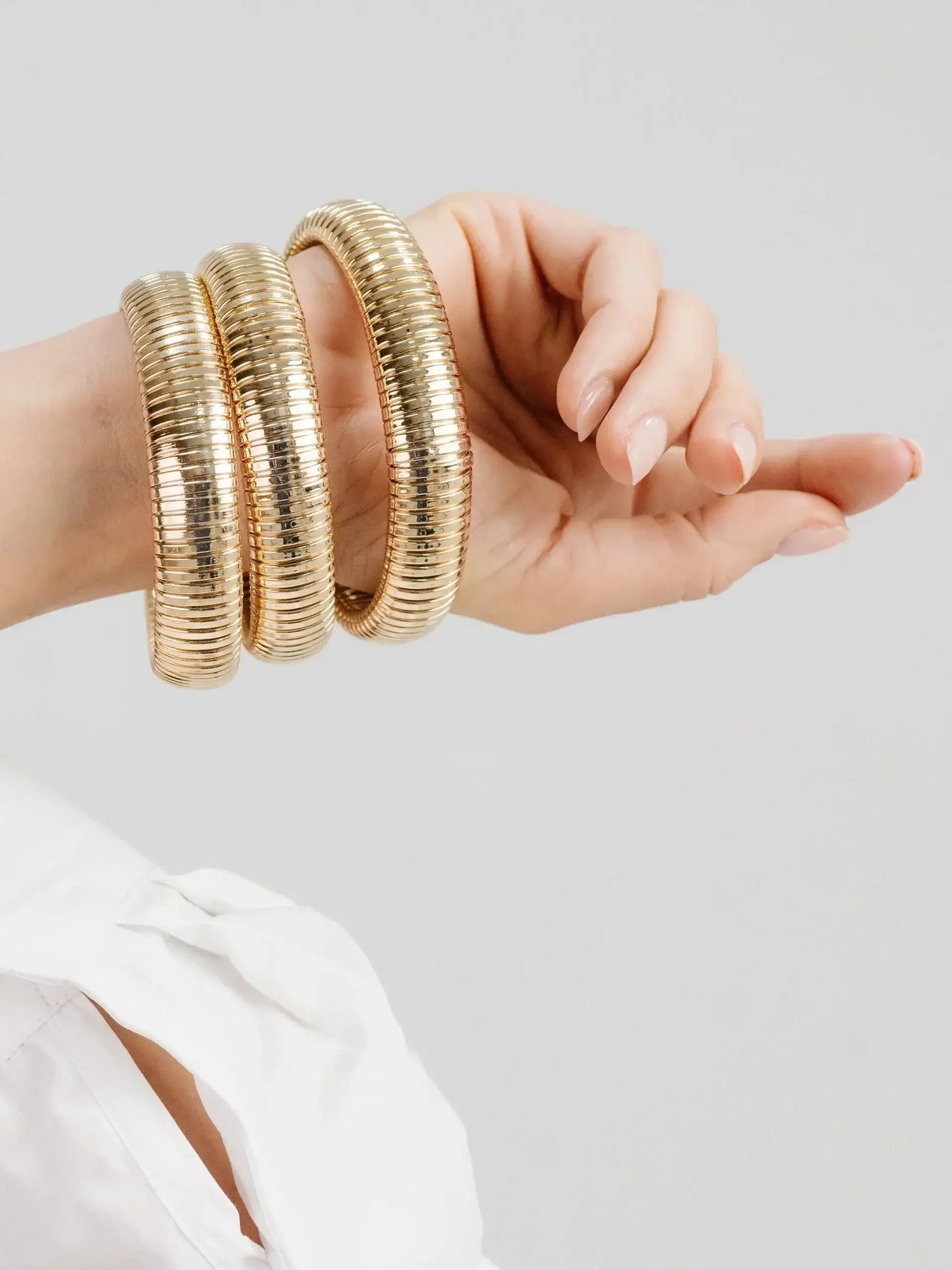 A hand adorned with three shiny gold bangles is raised, showcasing their smooth, ridged design. The background is neutral, emphasizing the jewelry and the hand's elegant posture.