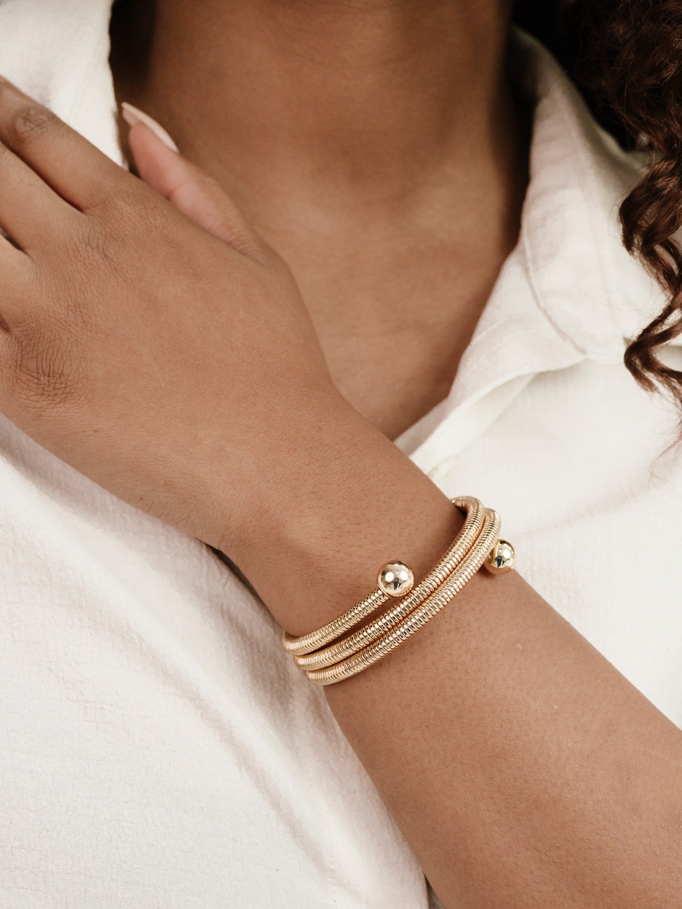 A hand rests on a shoulder, adorned with a multi-stranded gold bracelet featuring gleaming beads. The individual wears a light-colored, textured shirt, set against a neutral background.