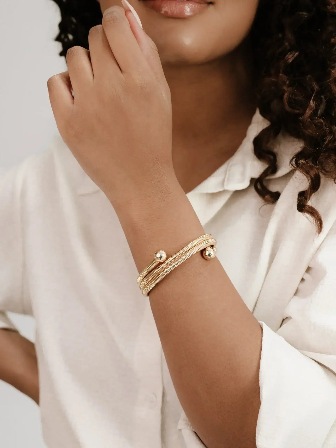 A hand with an elegant gold bracelet rests near the chin. The person wears a light-colored, button-up shirt, and soft lighting creates a serene atmosphere in the setting.