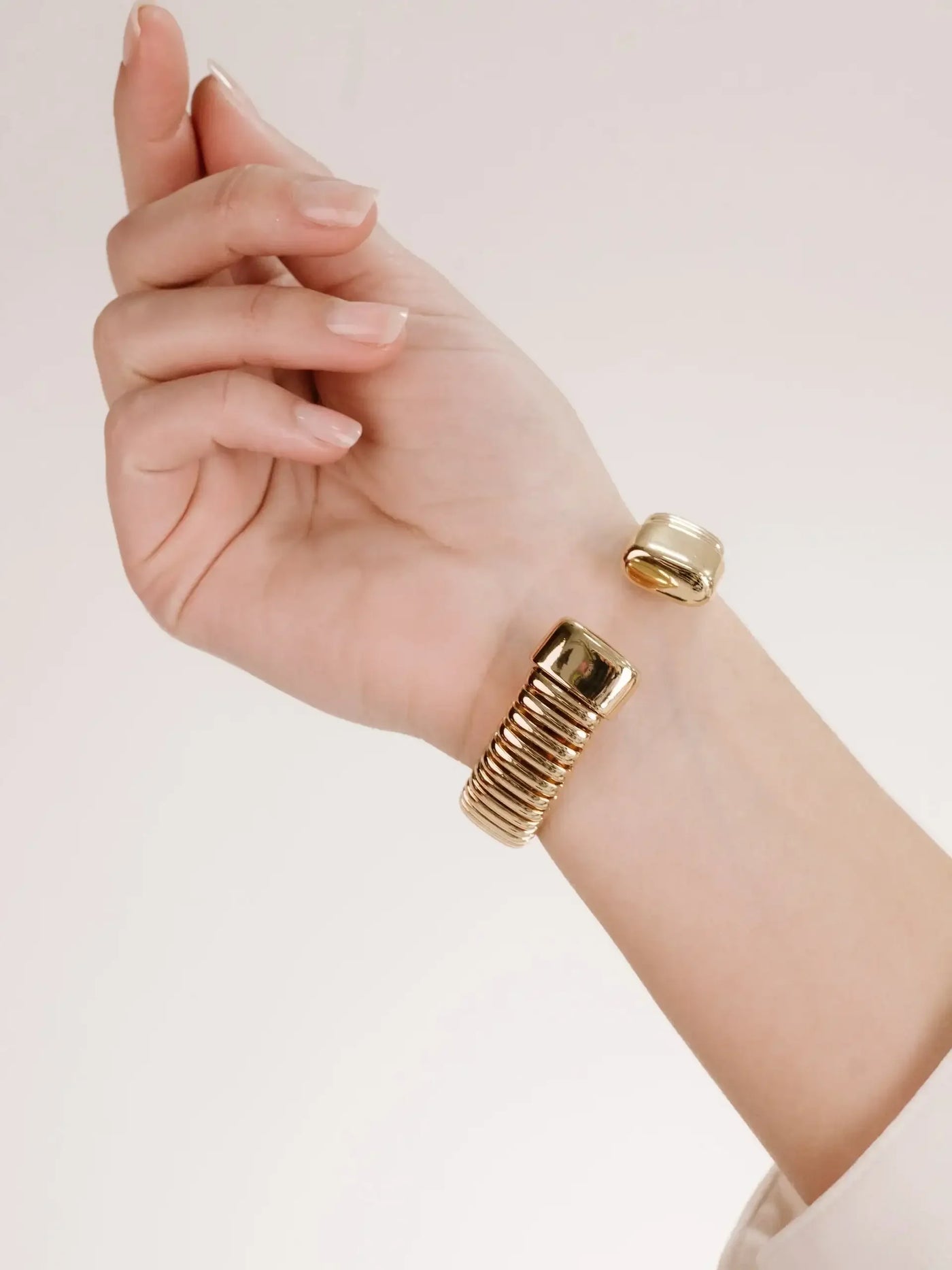A hand with neatly manicured nails displays a gold bangle bracelet featuring a ribbed design and a sleek, shiny surface, set against a neutral background.