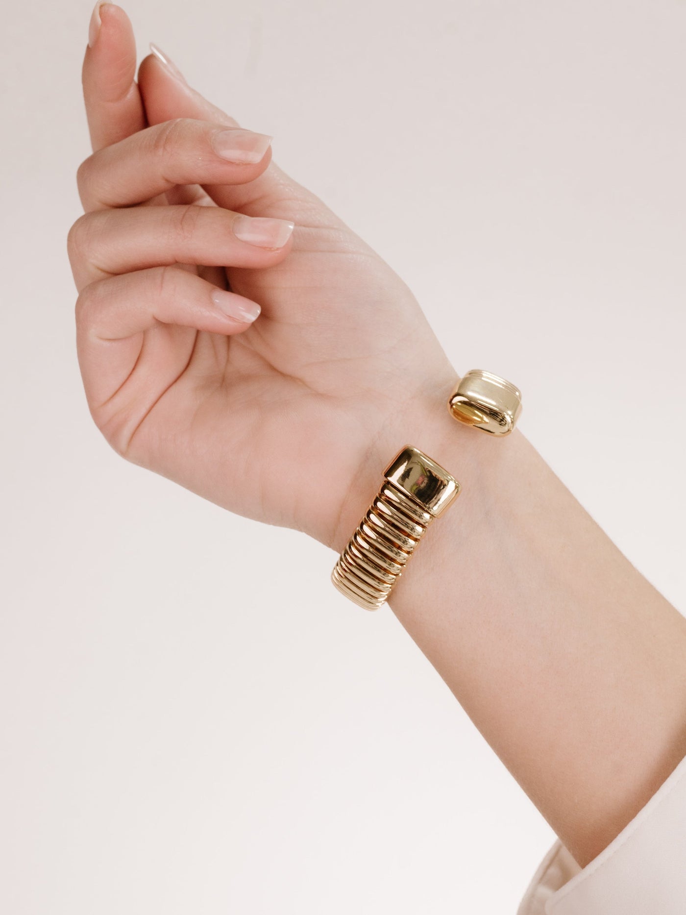 A hand with neatly manicured nails displays a gold bangle bracelet featuring a ribbed design and a sleek, shiny surface, set against a neutral background.