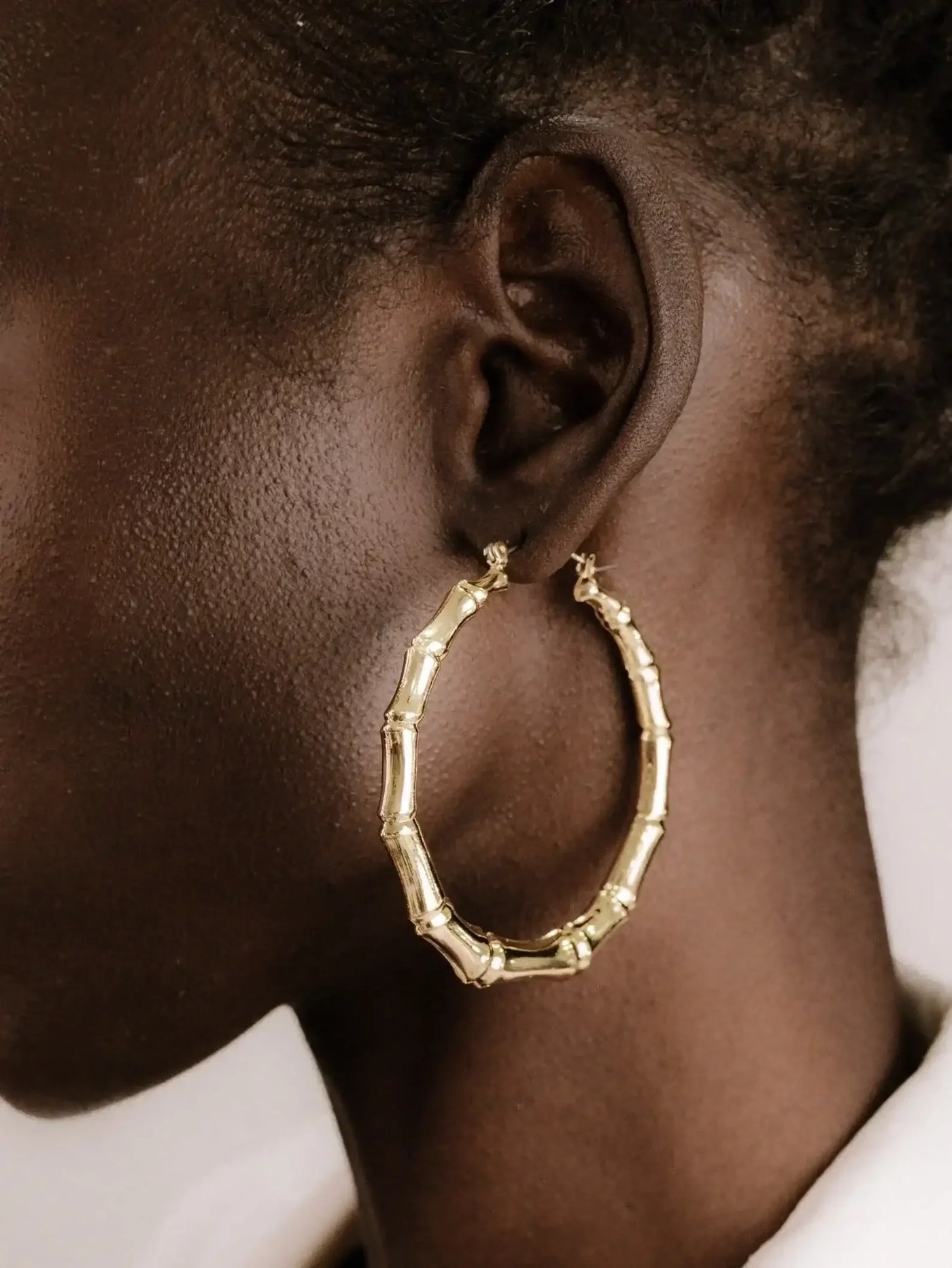 A large, gold bamboo hoop earring hangs from the earlobe of a person with dark skin. The background is neutral, highlighting the earring's design.