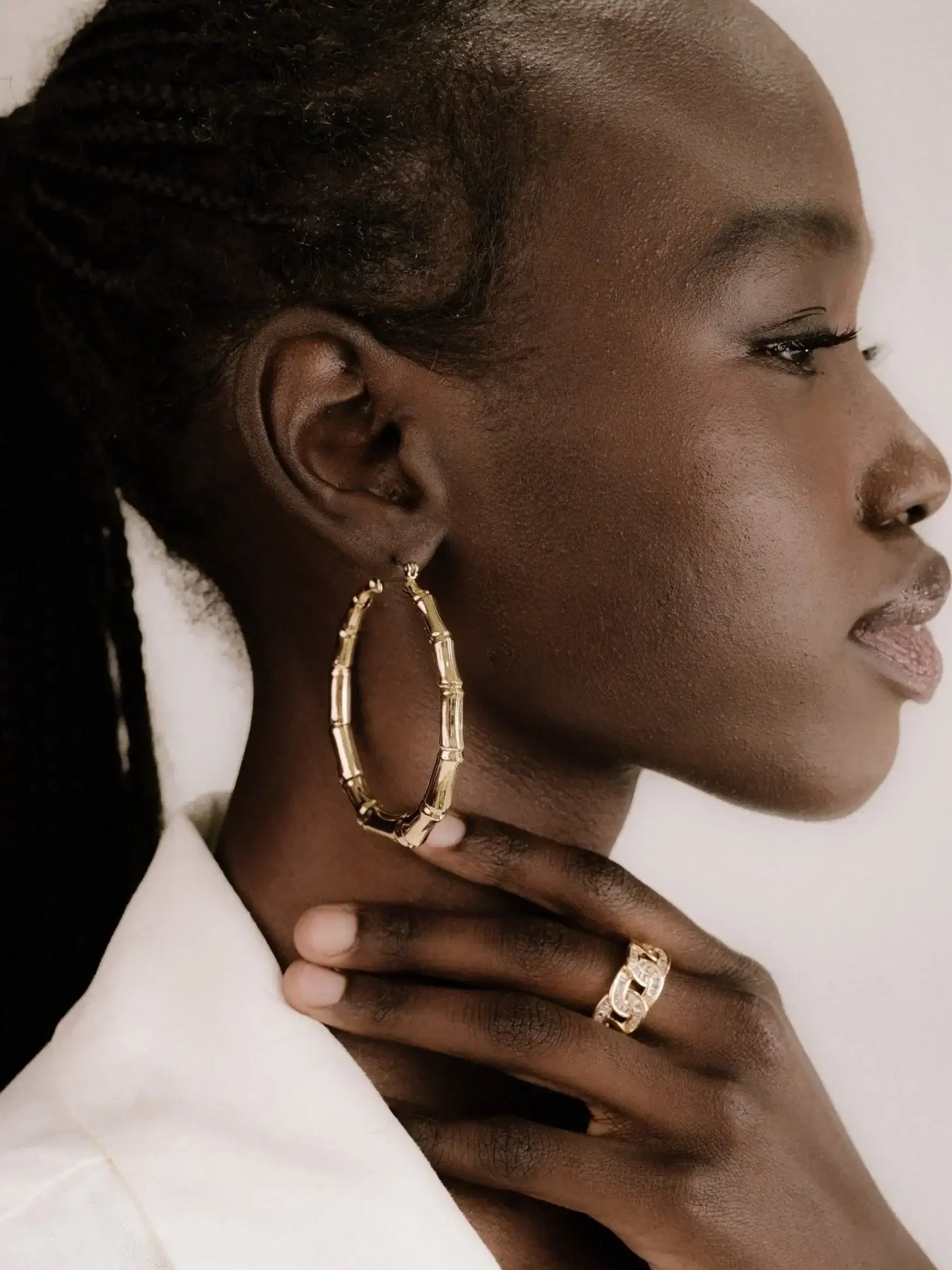 A woman wears large gold hoop earrings and a ring, gently touching her neck with one hand. The background is neutral, highlighting her accessories and smooth facial features.