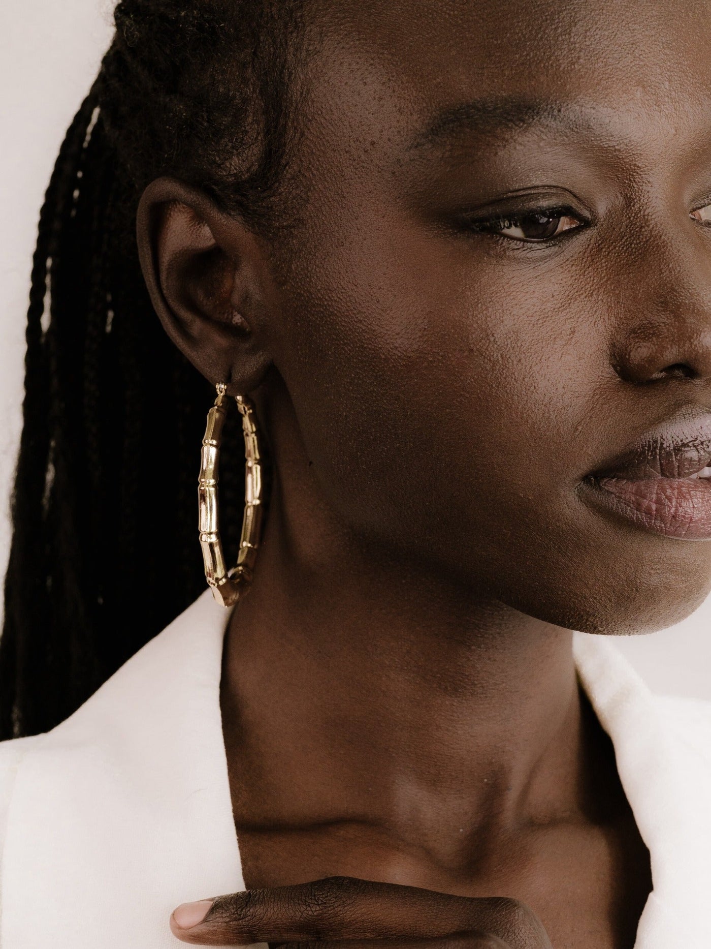 A close-up of a dark-skinned woman wearing large, golden hoop earrings, gently touching her collarbone with one hand while looking thoughtfully to the side against a soft, neutral background.