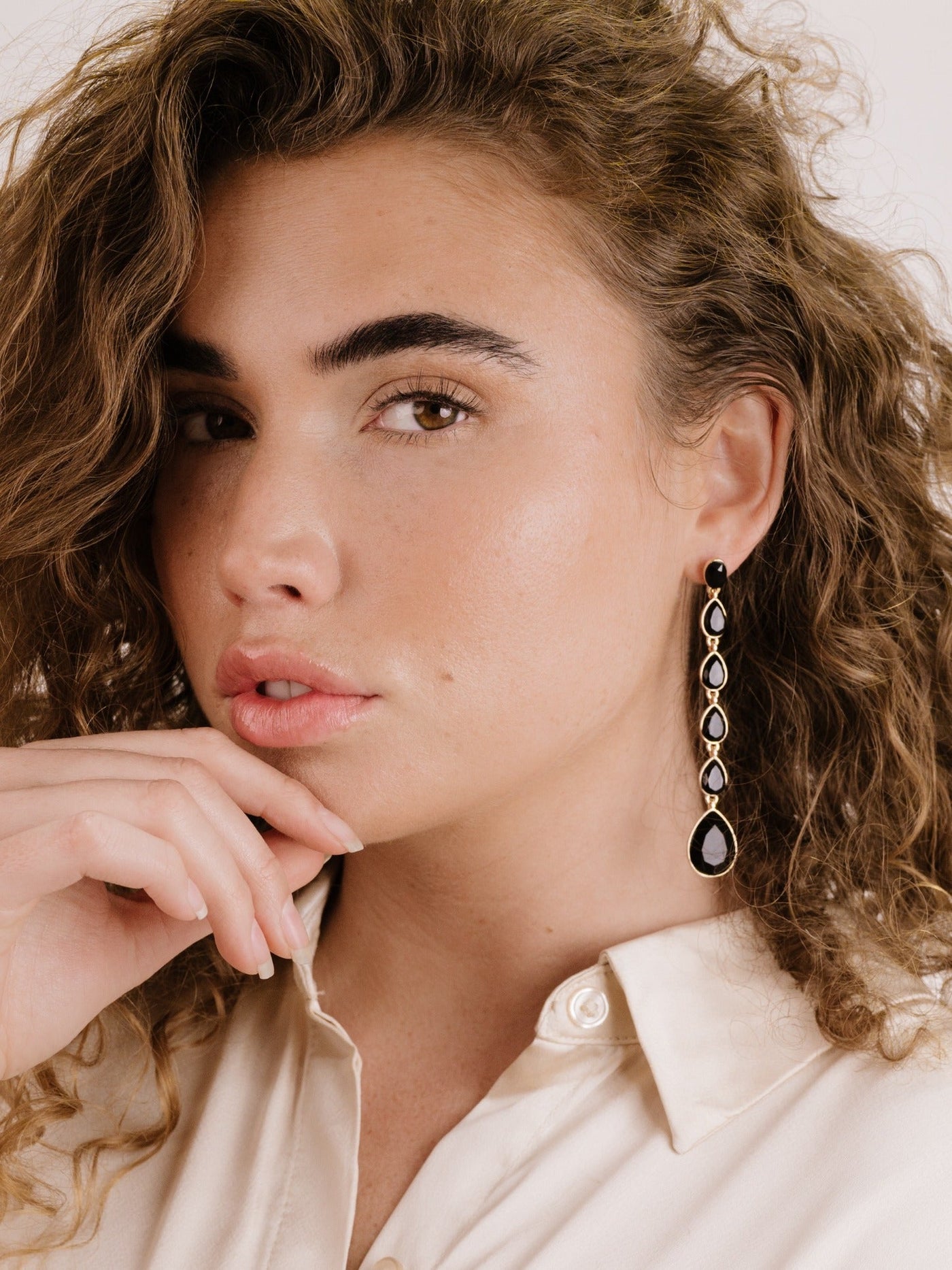 A woman with curly hair wears an elegant, long earring featuring black and gold elements. She rests her hand thoughtfully on her chin, set against a neutral background.