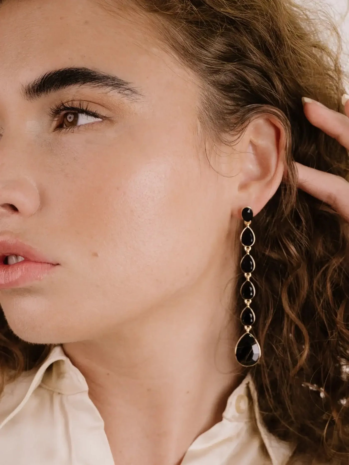 A close-up of a woman with curly hair, showcasing a long black earring. She gently touches her hair, with her face partially turned to the side against a neutral background.