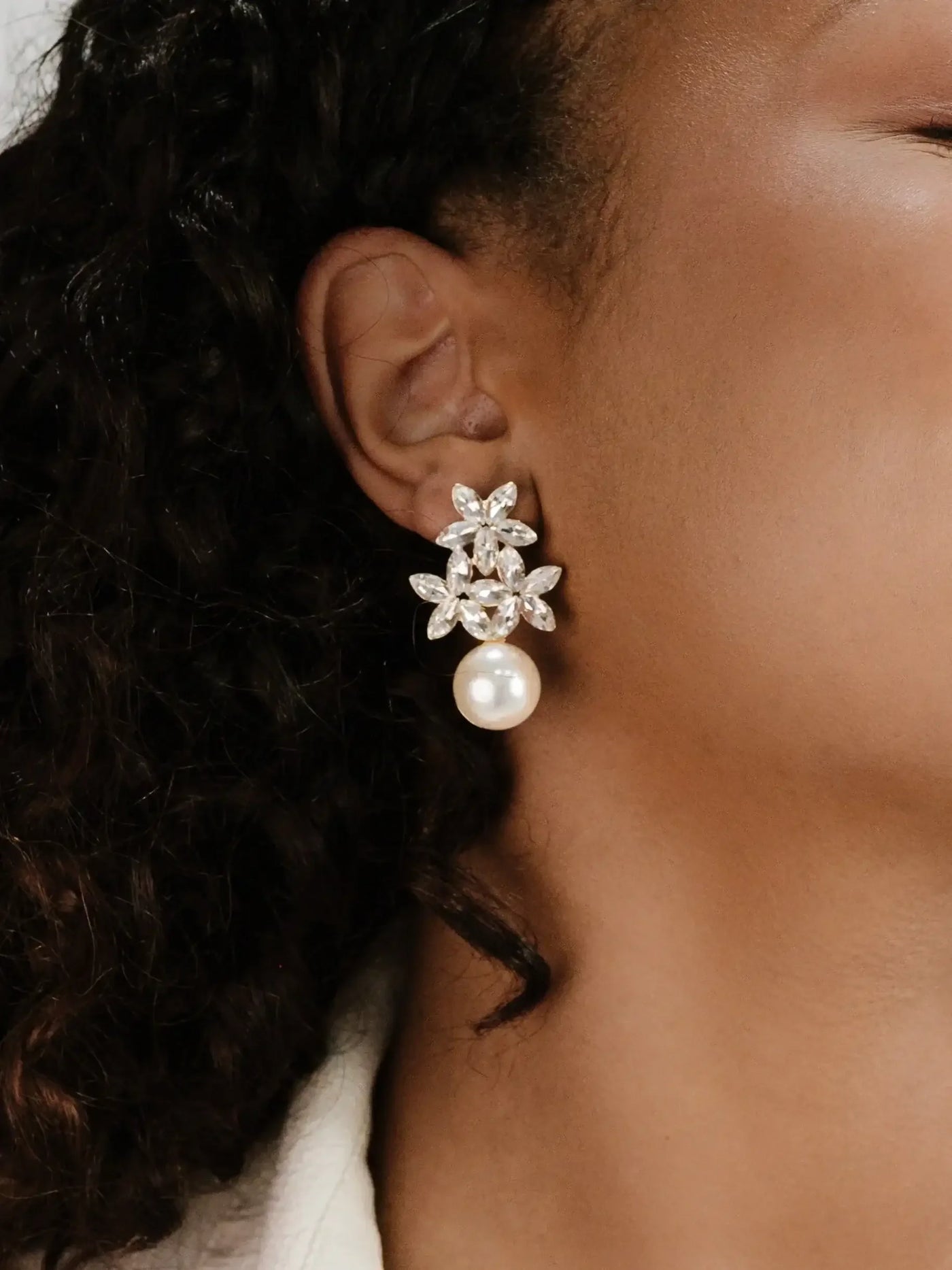 A floral-designed earring featuring clear gemstones and a large pearl hangs from the ear of a person with curly hair, set against a softly lit, neutral background.