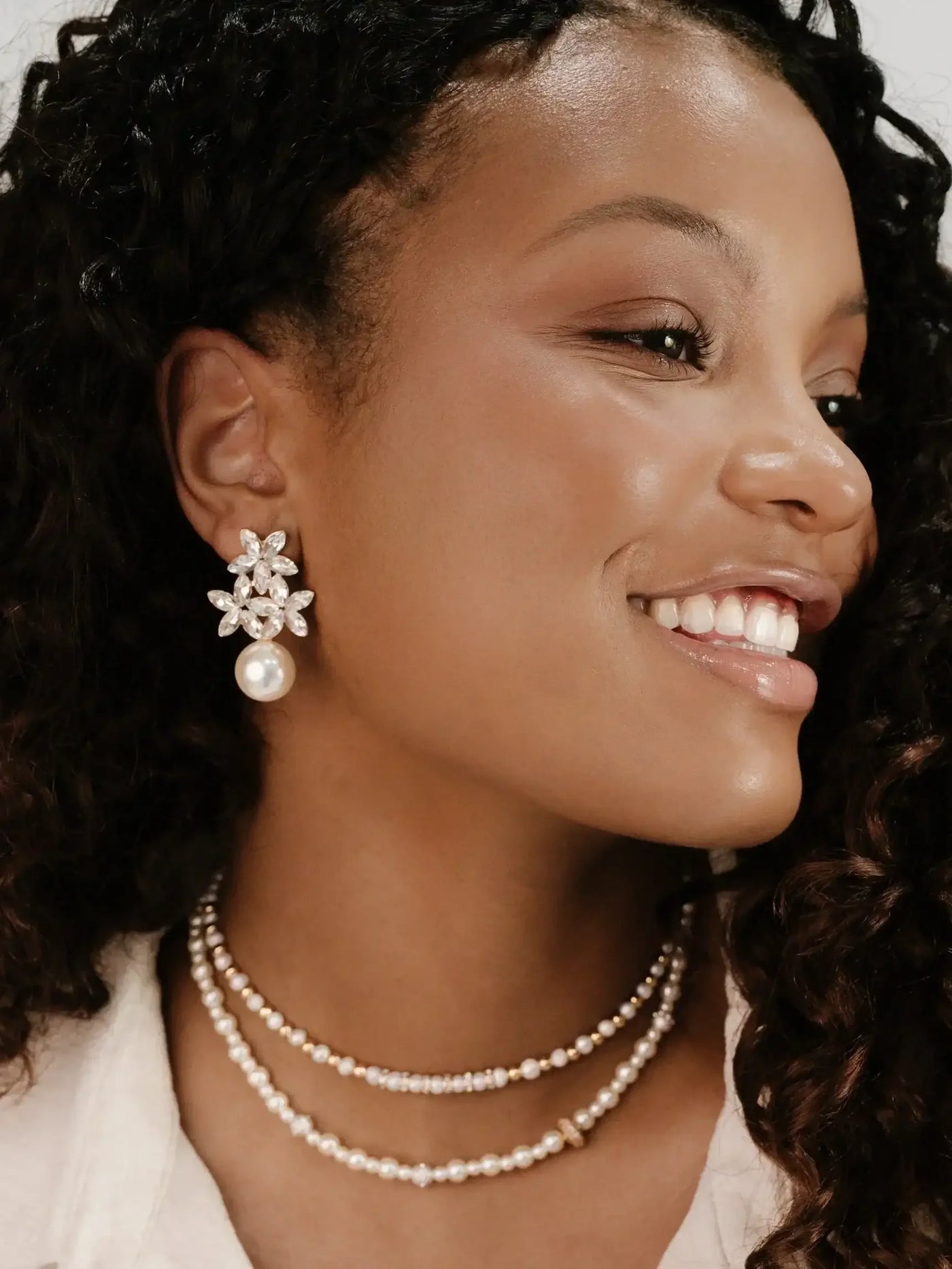 A woman with curly hair smiles, showcasing ornate earrings with floral designs and a pearl. She wears multiple delicate pearl necklaces against a softly illuminated background.