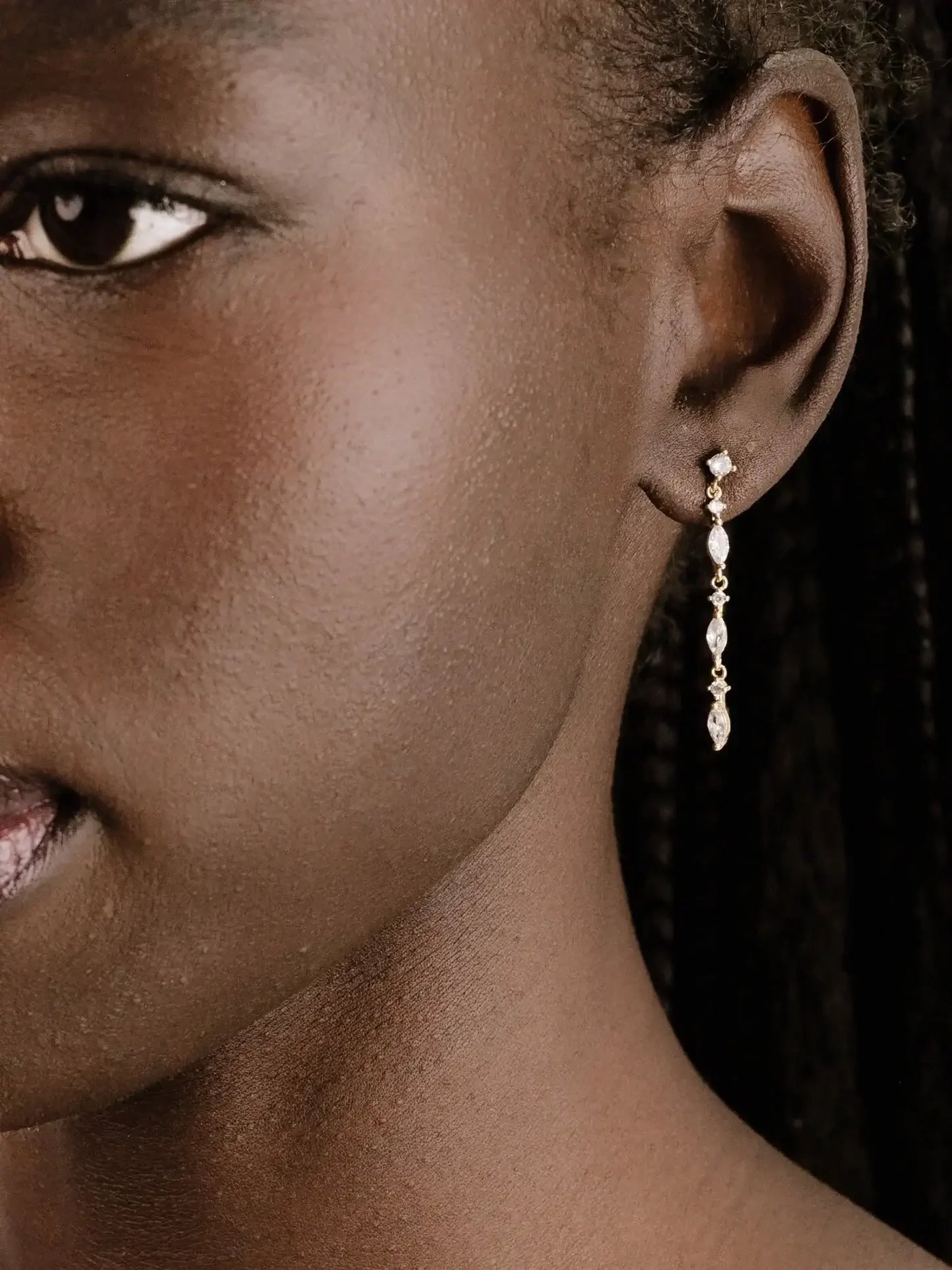 A close-up view of an earlobe showcasing a delicate, dangling earring with a gemstone, highlighting smooth, dark skin and intricately braided hair in the background.