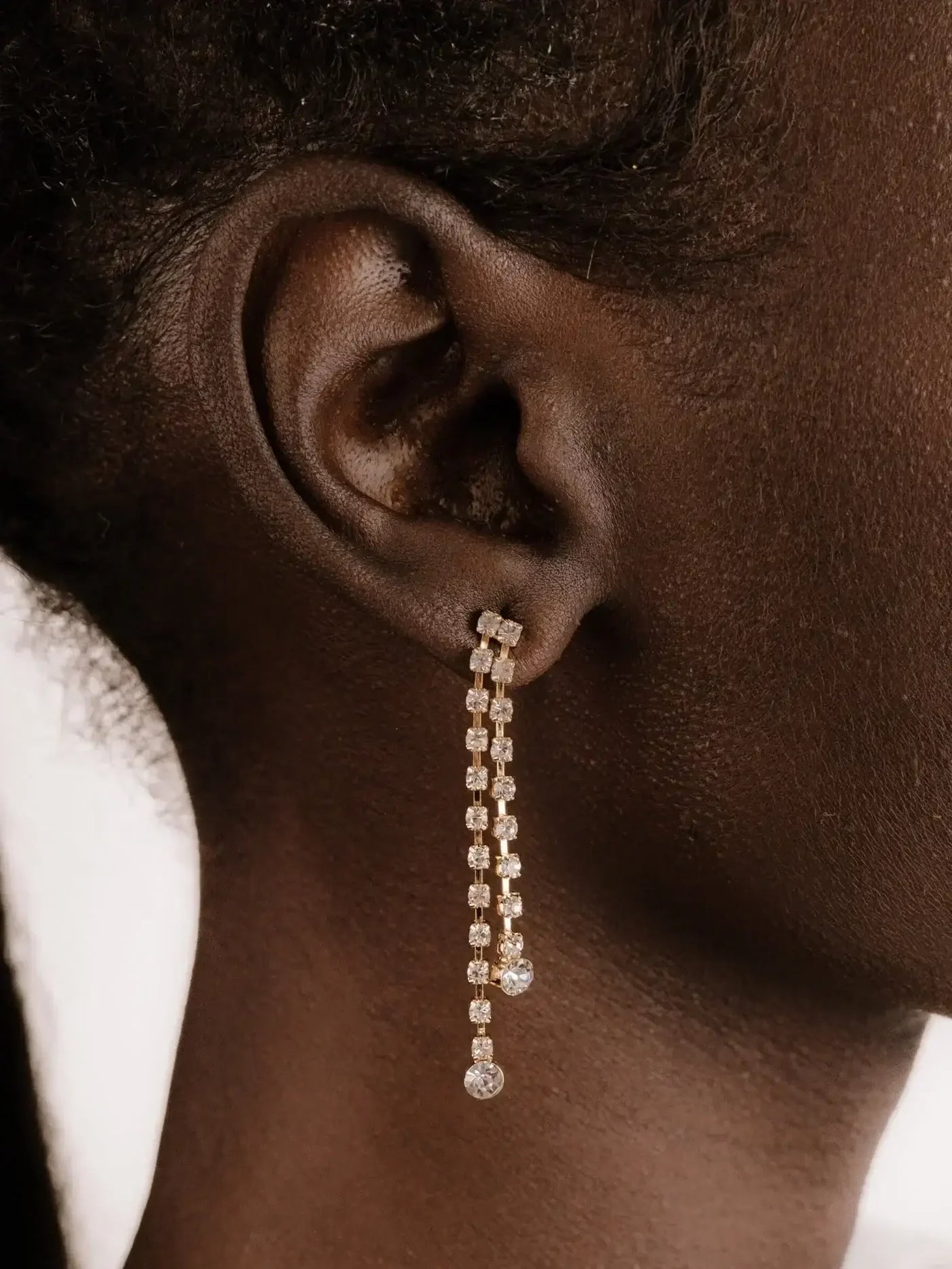 A long, elegant earring featuring sparkling crystals hangs from a pierced ear, accentuating the skin tone and texture of the model's neck and nearby hair.