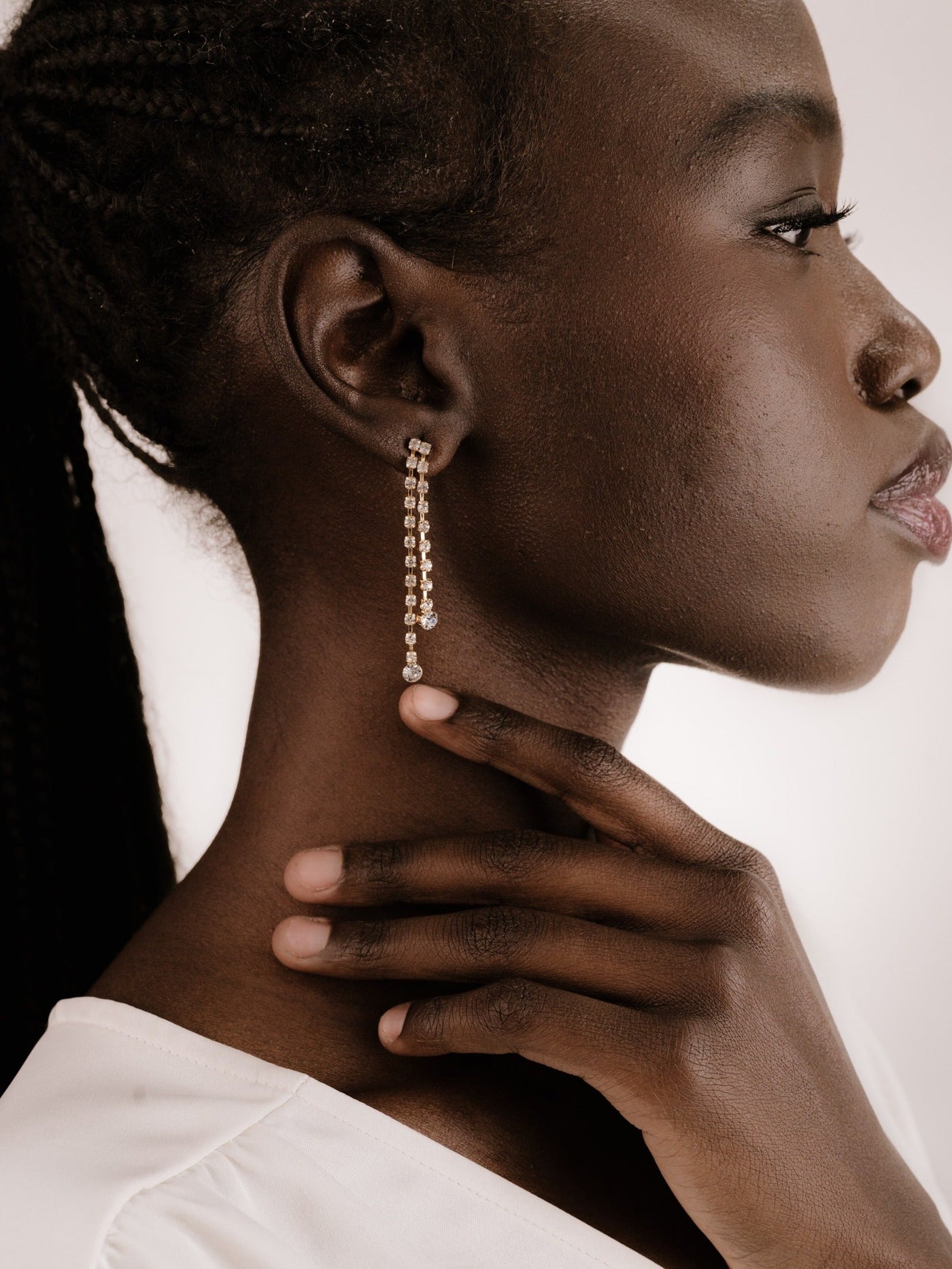 A profile of a woman with braided hair touches her neck, adorned with a dangling earring featuring small, shiny beads. The background is neutral and softly lit.