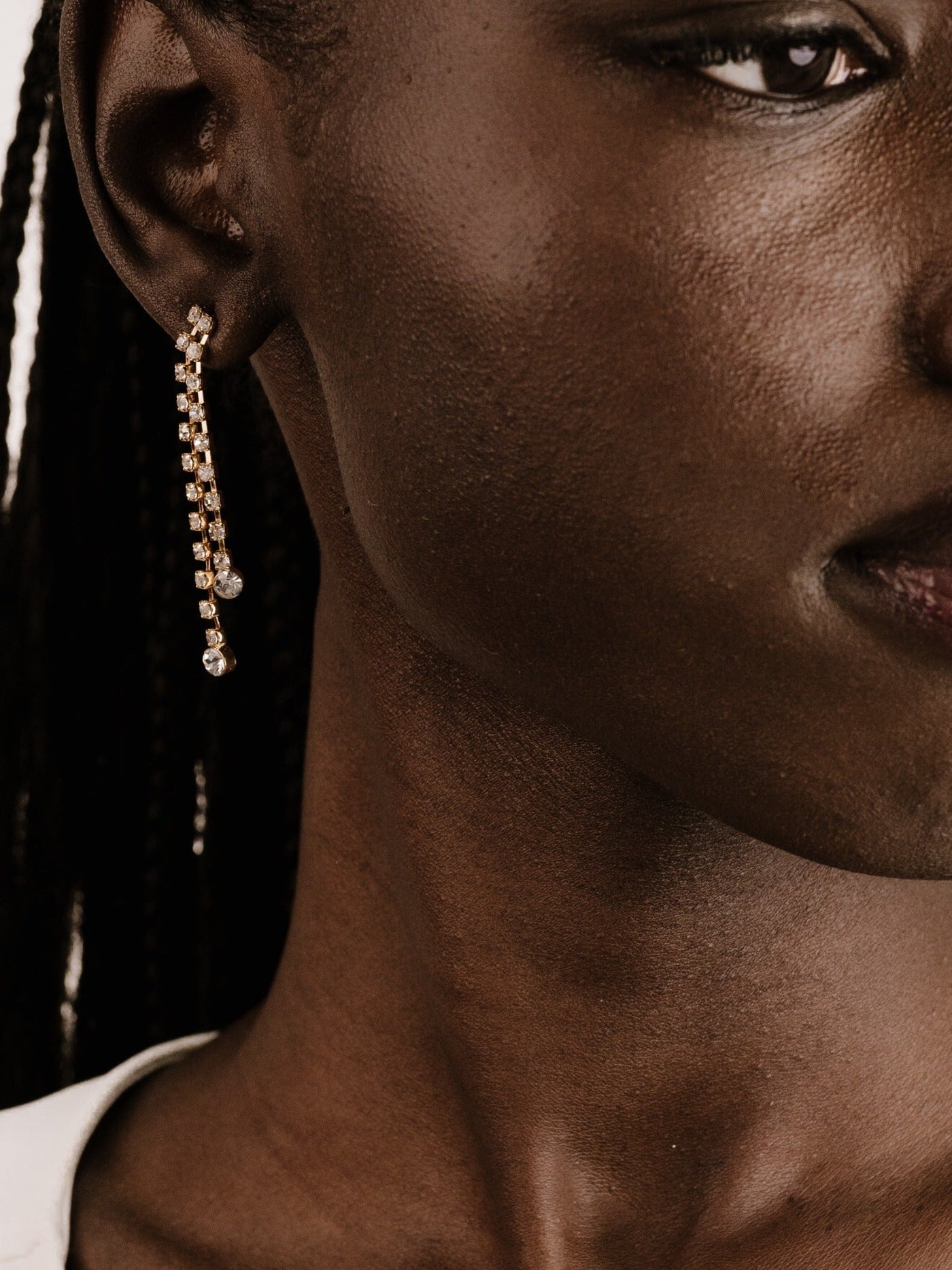 A close-up of an ear adorned with a long, sparkling earring. The skin tone is rich and textured, with braided hair visible in the background against a neutral setting.
